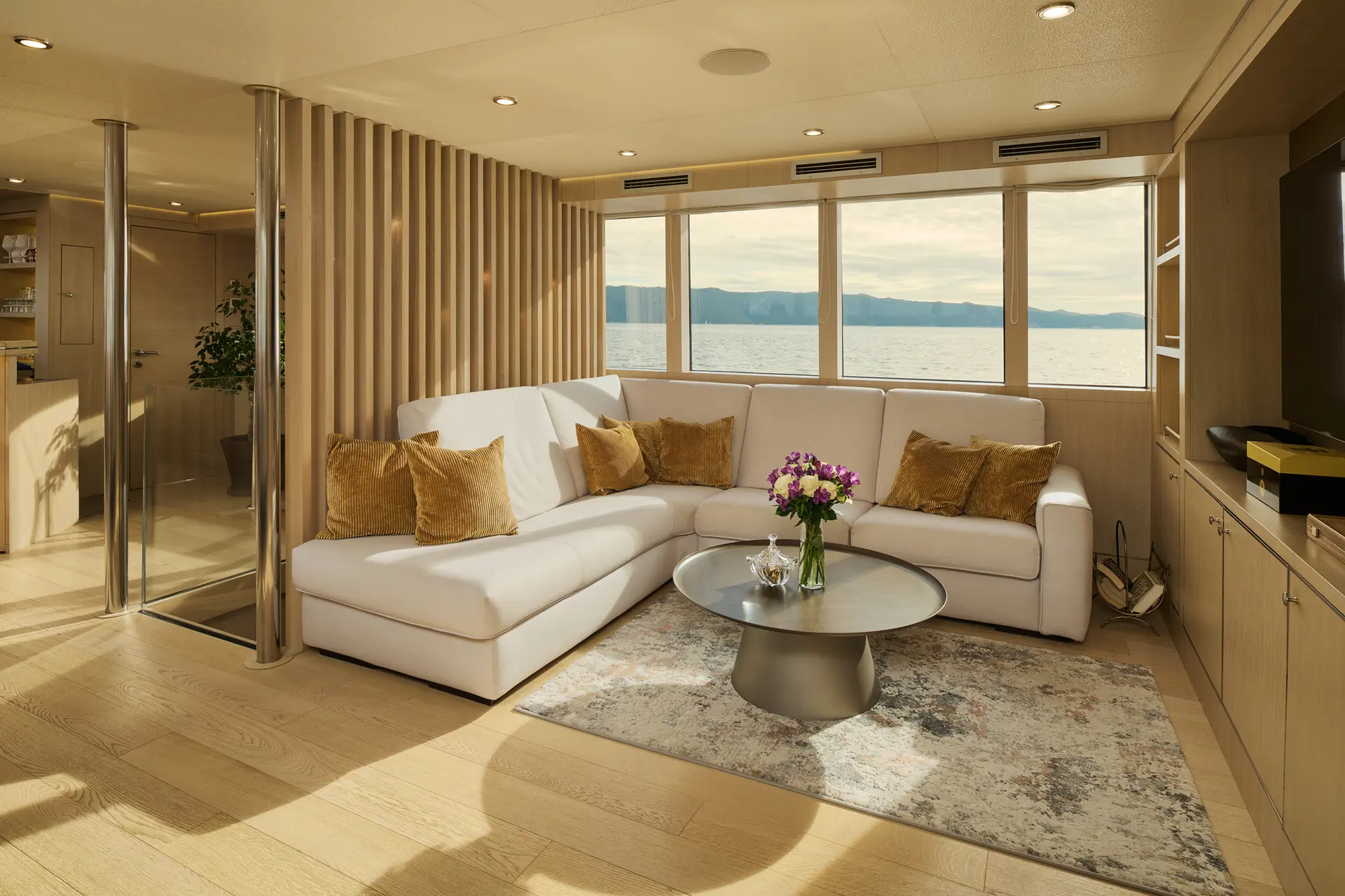 Modern yacht living room with large windows overlooking water, a white sectional sofa with gold pillows, a round coffee table holding a flower vase, and light wood floors and walls with a scenic outdoor view.