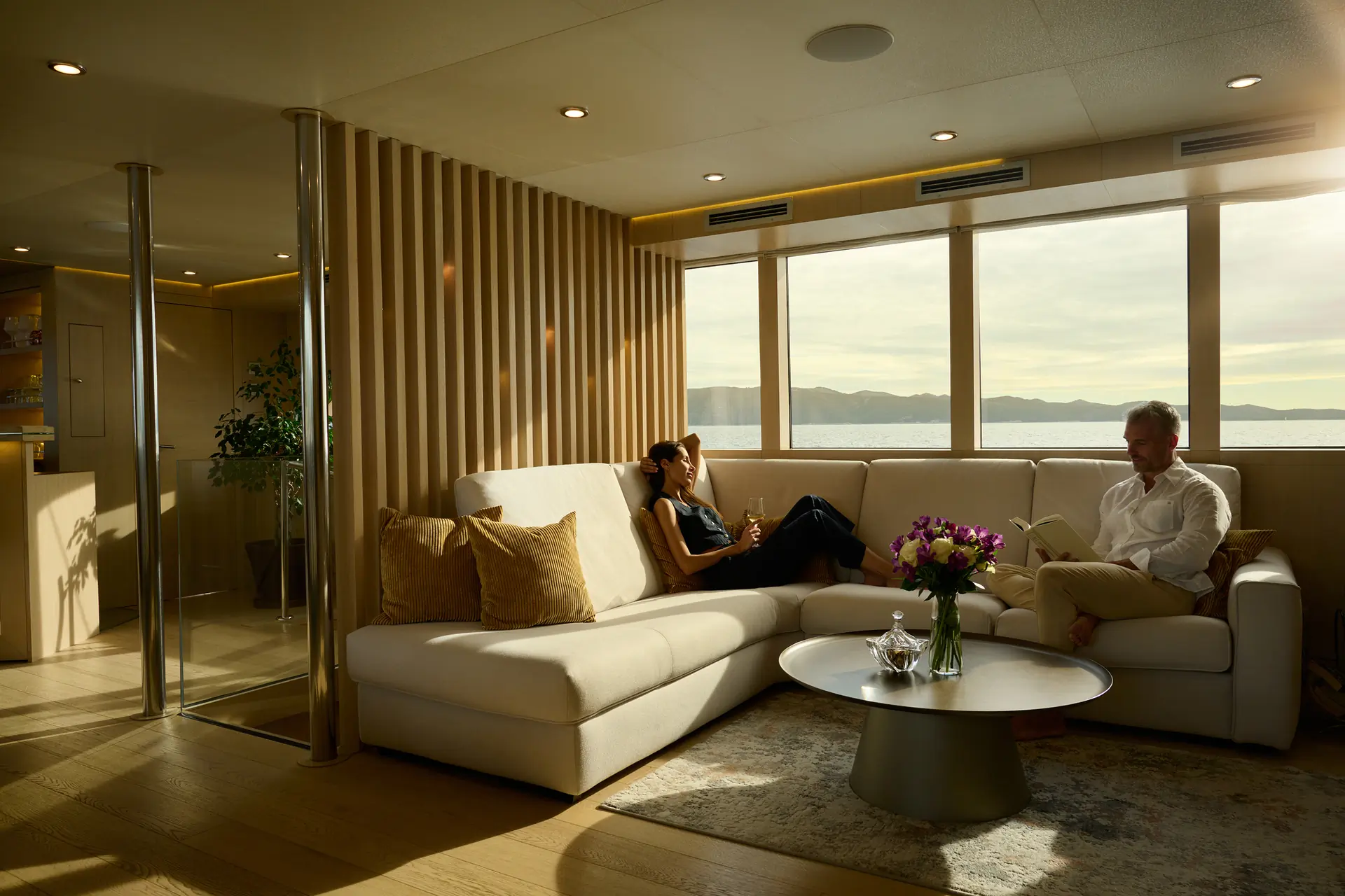 A woman and a man relax on a white sectional sofa in a modern, sunlit living room with large windows, wooden accents, and a round table with flowers, overlooking a scenic water view.