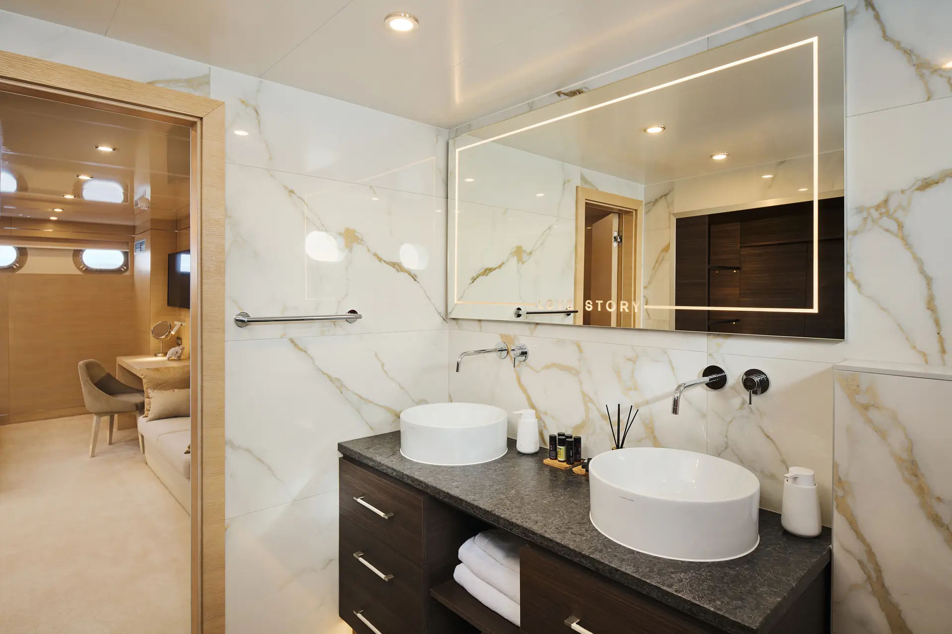 Modern bathroom with marble walls, double sinks on a dark countertop, large illuminated mirror, and wooden cabinets. Through the open door, a glimpse of a beige room with sofas and round windows is visible.
