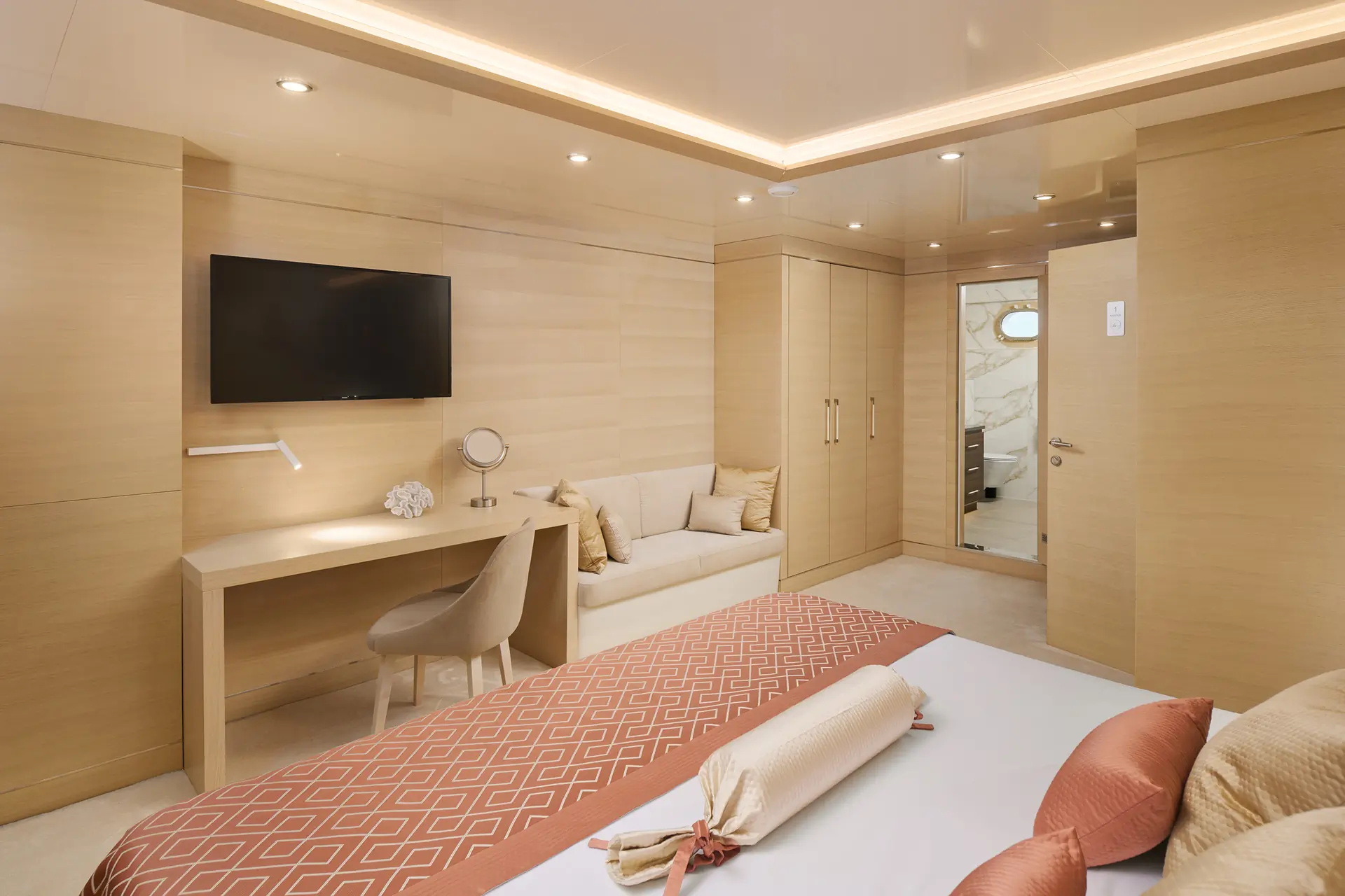 Modern bedroom with light wood walls, a double bed with pink and cream bedding, a wall-mounted TV, desk with chair, small sofa, and an open door showing a bathroom with marble walls.