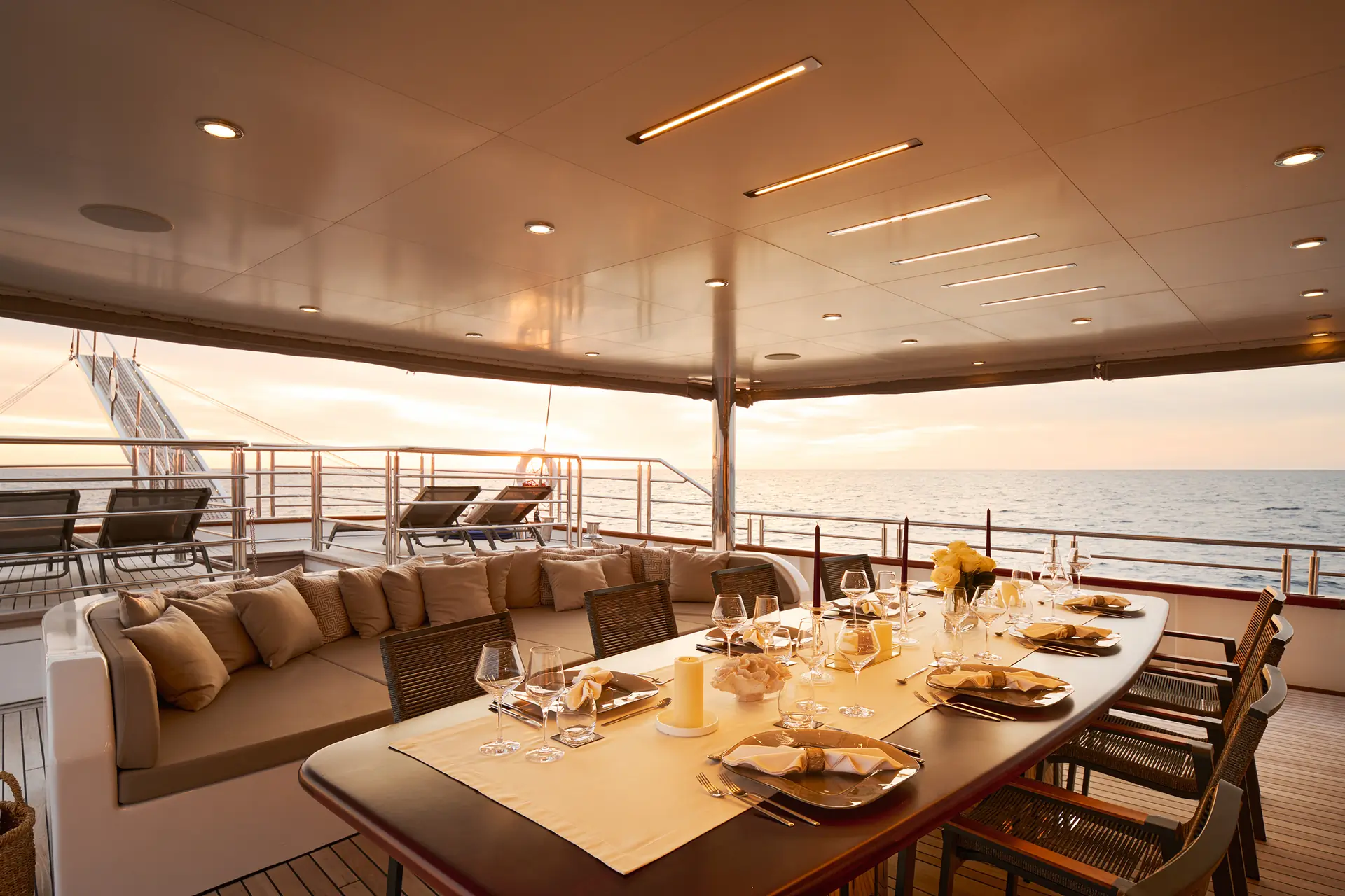 Elegant outdoor dining area on a yacht at sunset, featuring a set table with plates, glasses, and flowers, cushioned seating to the left, and views of the calm ocean and sky in the background.