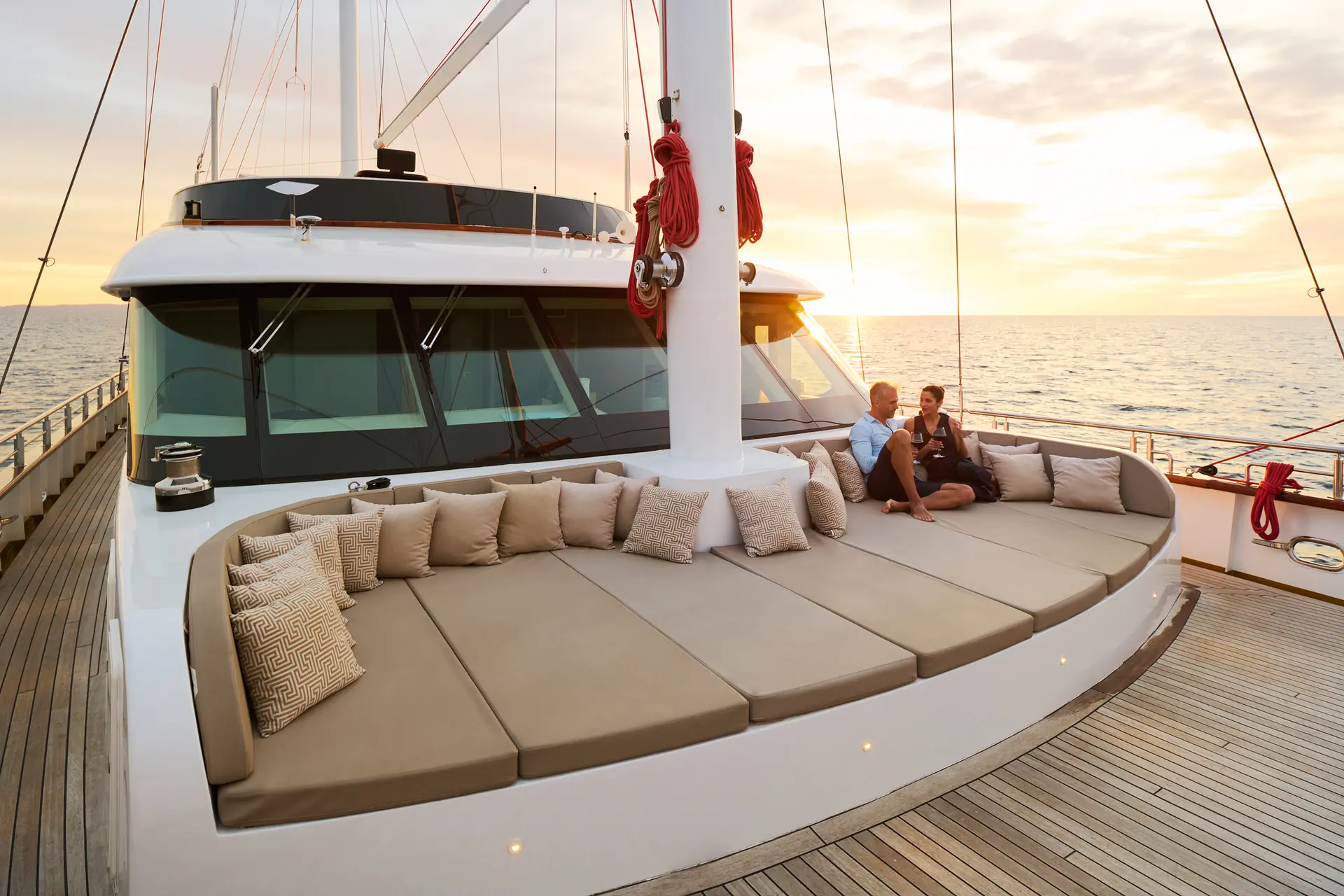 A couple sits on a large cushioned lounge area at the front of a yacht, enjoying the sunset over a calm sea. Pillows are arranged around the lounge, and the atmosphere is peaceful and relaxing.