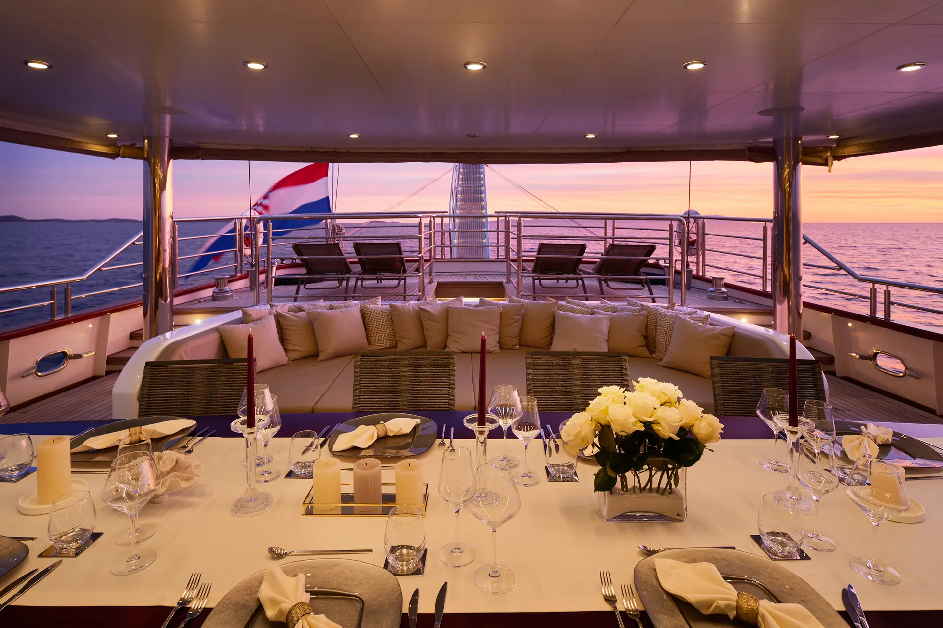 Elegant yacht dining area at sunset, with a set table, cushioned lounge, and a national flag on the stern. The ocean and a colorful sky are visible in the background.