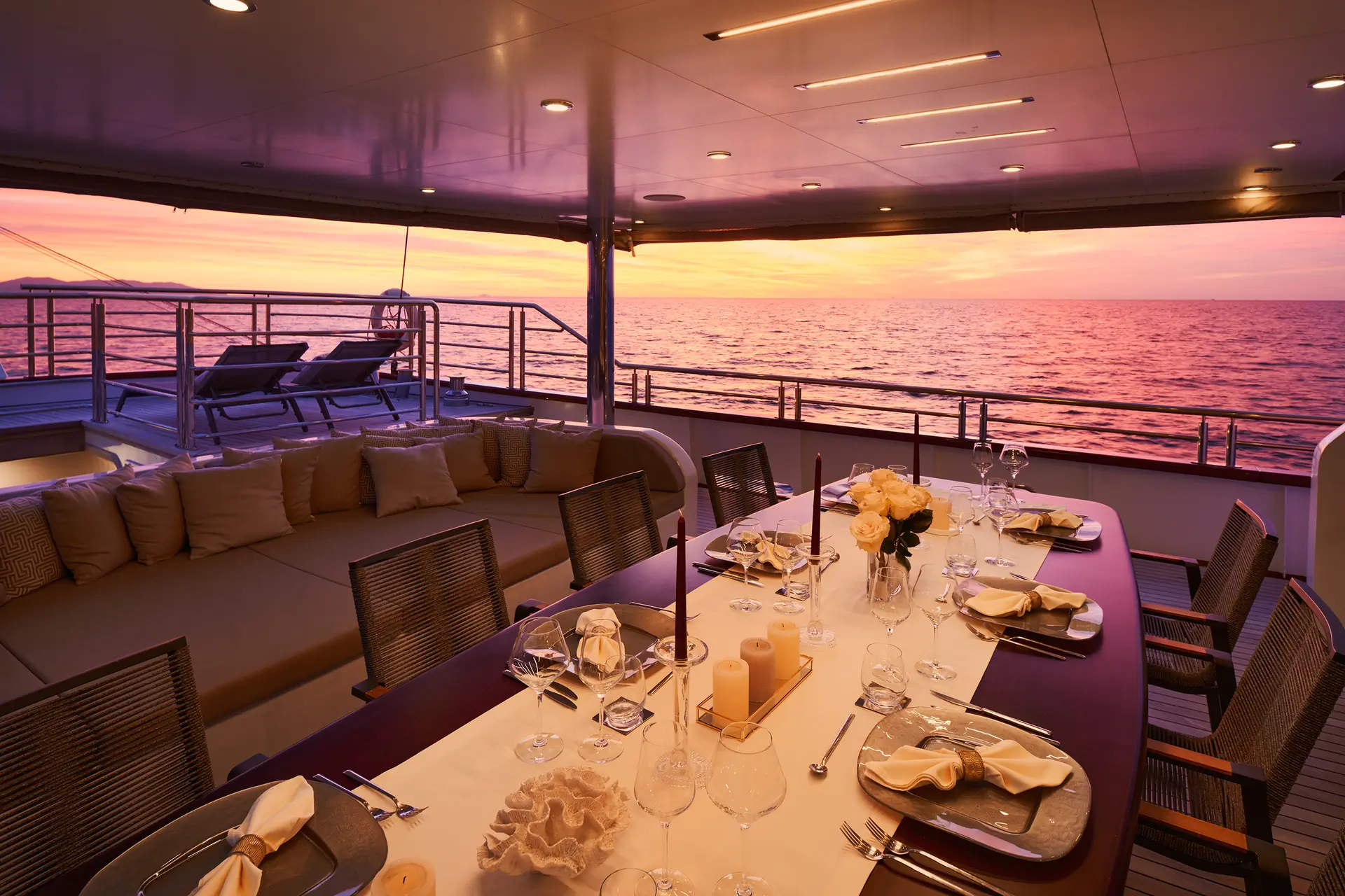 Elegant dining table set for dinner on a yacht deck, overlooking the ocean at sunset with a purple and orange sky. Lounge chairs and cushions are visible in the background.