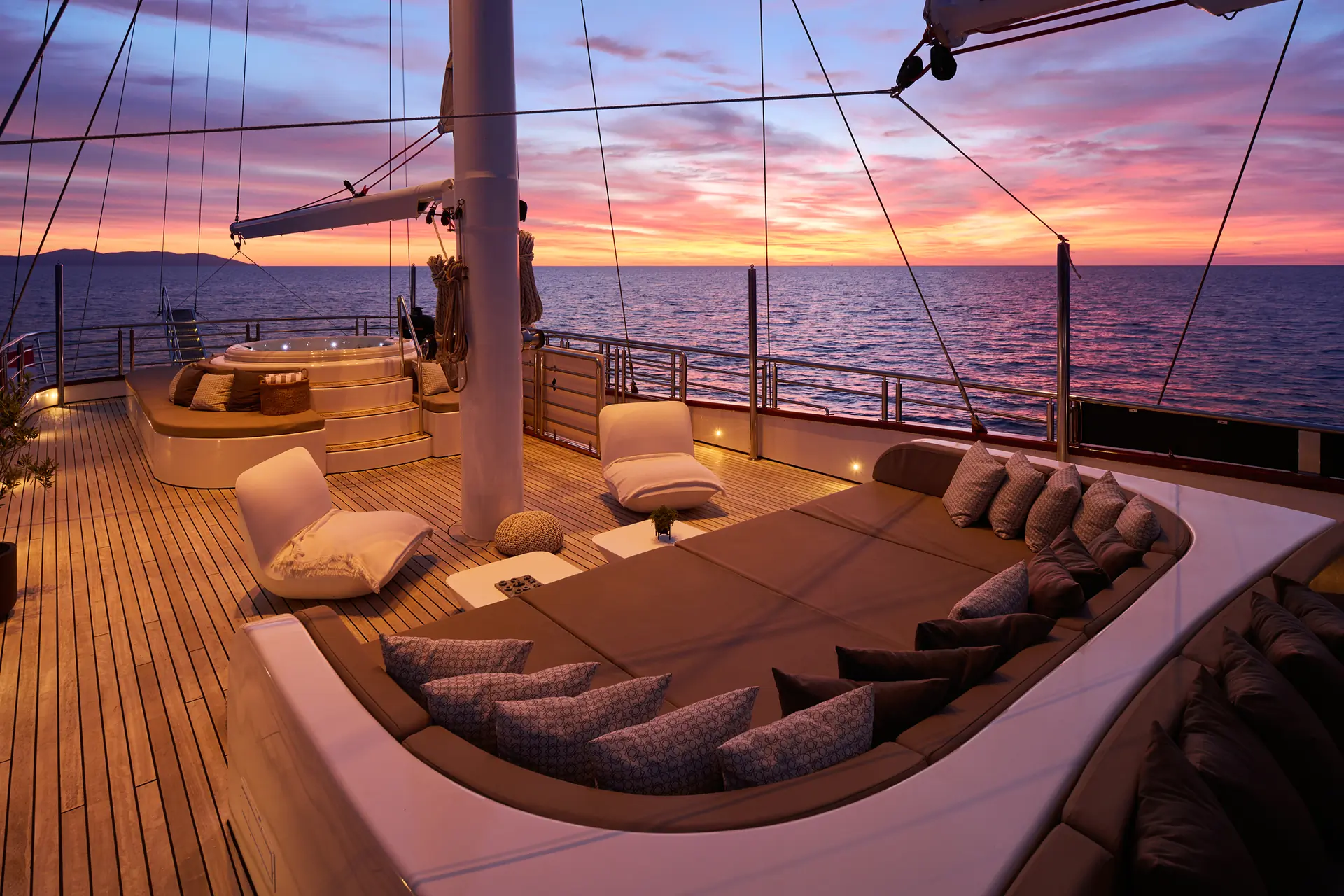 A luxurious yacht deck with cushioned seating and loungers overlooks the ocean at sunset, with vibrant orange and purple hues lighting up the sky and reflecting on the water.