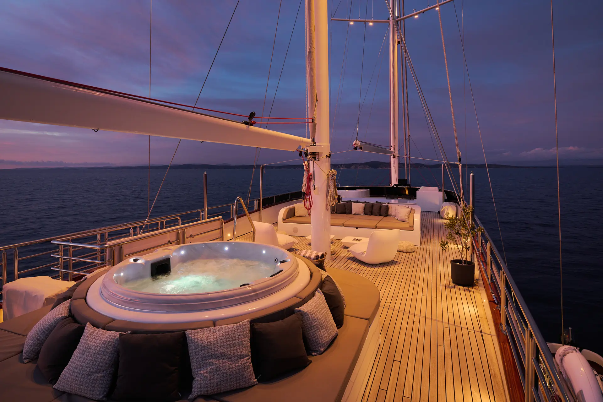 Luxury yacht deck at sunset with a round jacuzzi, cushion seating, modern furniture, and soft lighting, overlooking calm ocean waters and distant land on the horizon.