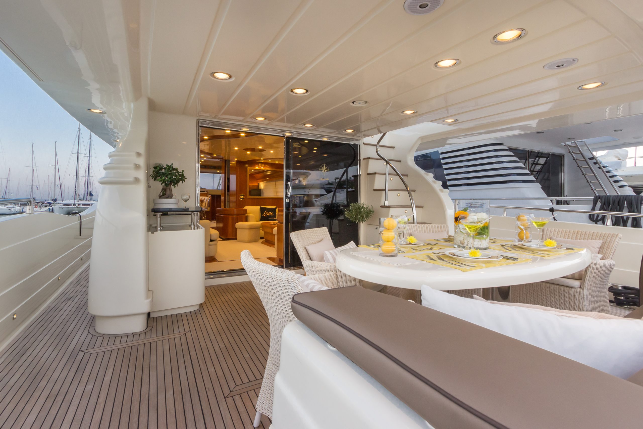 Luxurious yacht deck with a round dining table set for a meal, surrounded by wicker chairs and bench seating. Glass doors lead to an elegant interior, and marina boats are visible outside.