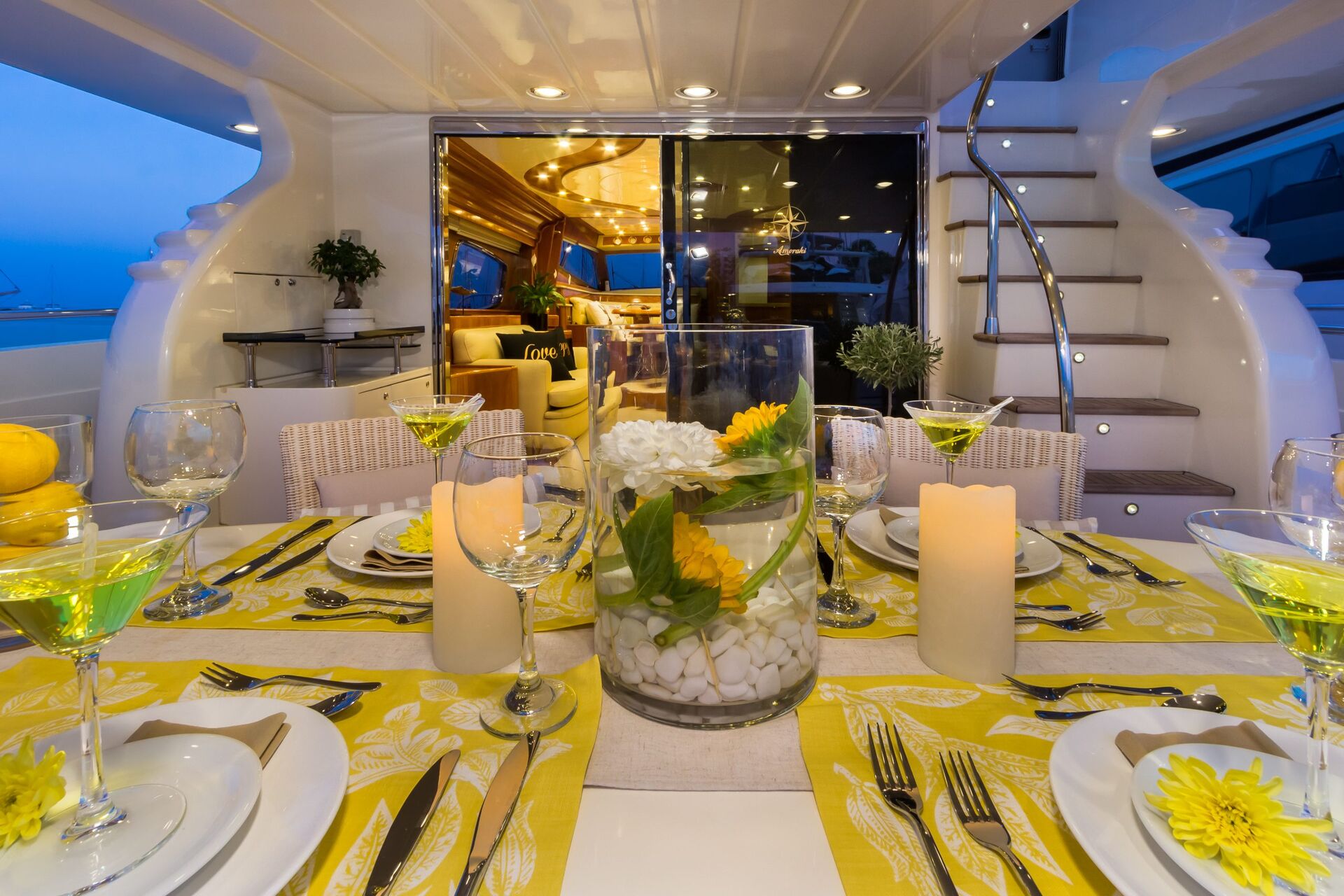 AMORAKI A luxurious yacht dining area set for dinner with yellow accents, green drinks, white plates, candles, and a floral centerpiece. The yacht’s elegant interior is visible through glass doors; spiral stairs are on the right.