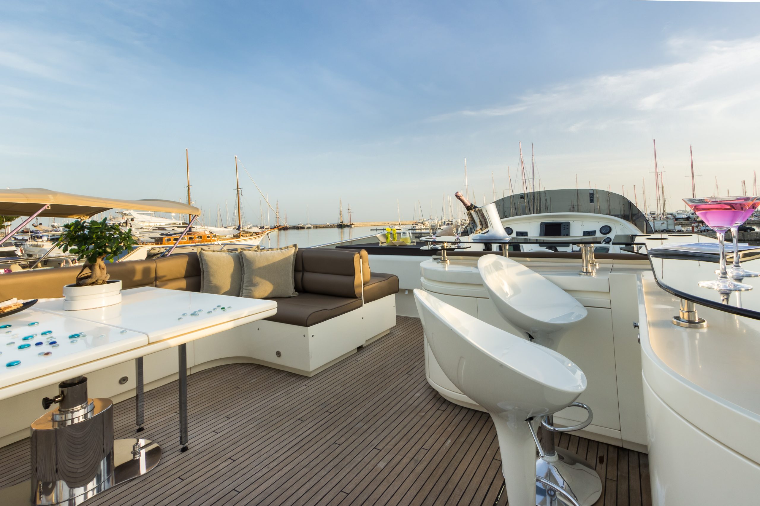 Open deck of a luxury yacht with white modern furniture, lounge seating, bar stools, drinks on the counter, and a view of a marina with several docked boats under a clear blue sky.