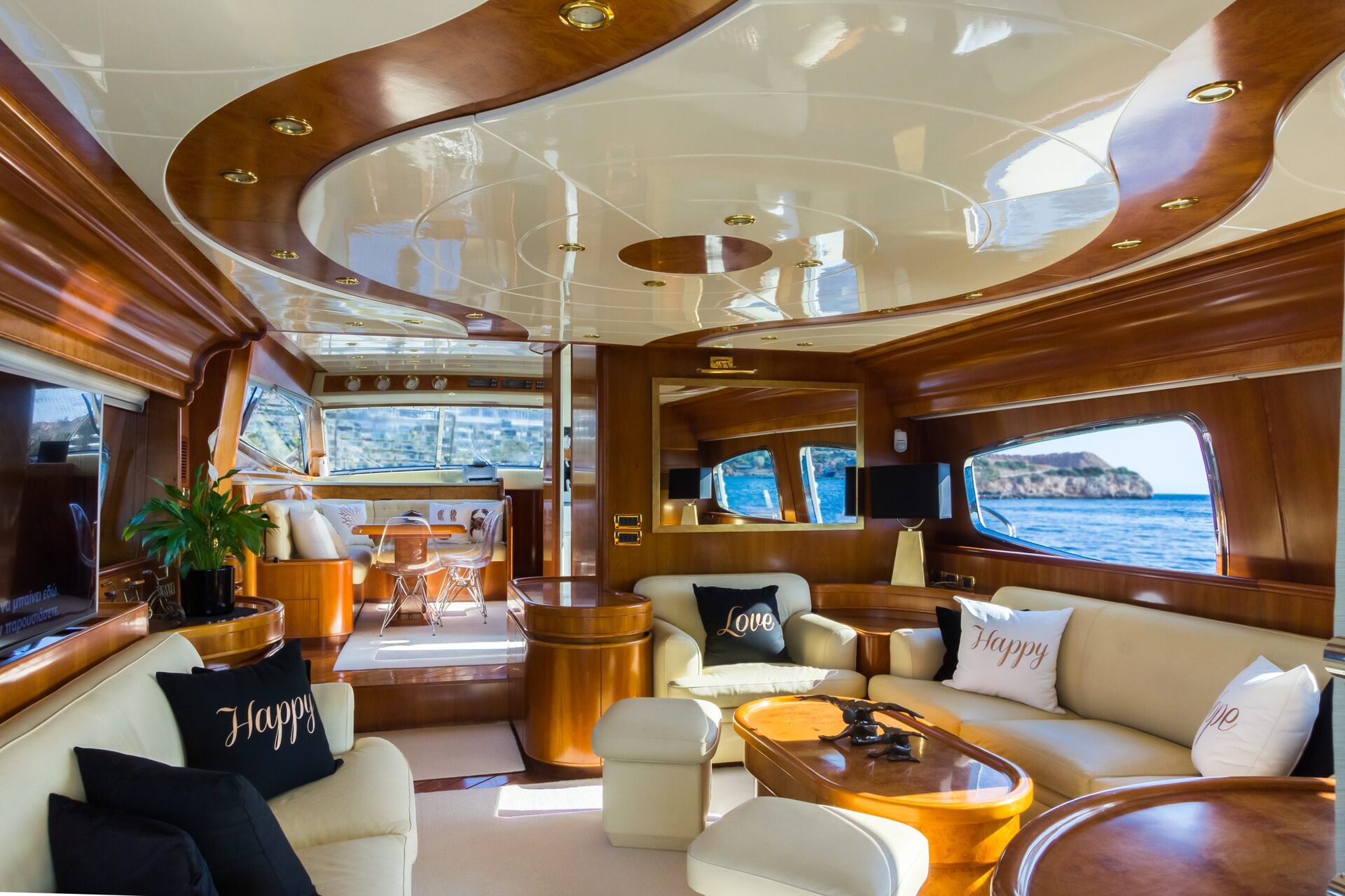 Luxurious yacht interior with polished wood, cream sofas, and decorative pillows reading “Love” and “Happy.” Large windows offer views of the ocean and cliffs; sunlight fills the bright, elegant space.
