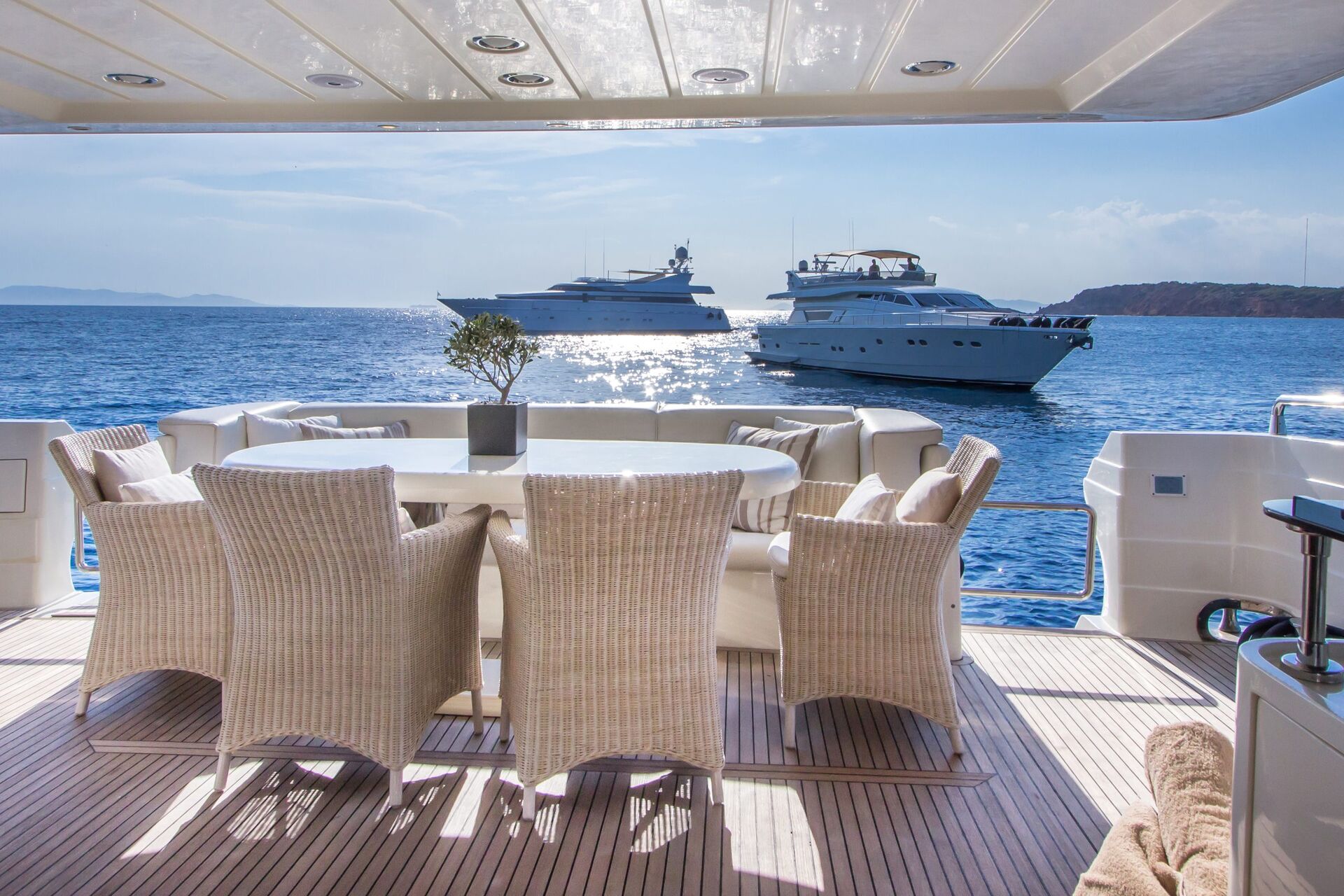 A luxury yacht deck with a round dining table and wicker chairs overlooks calm blue water, with two other yachts anchored nearby under a sunny sky.