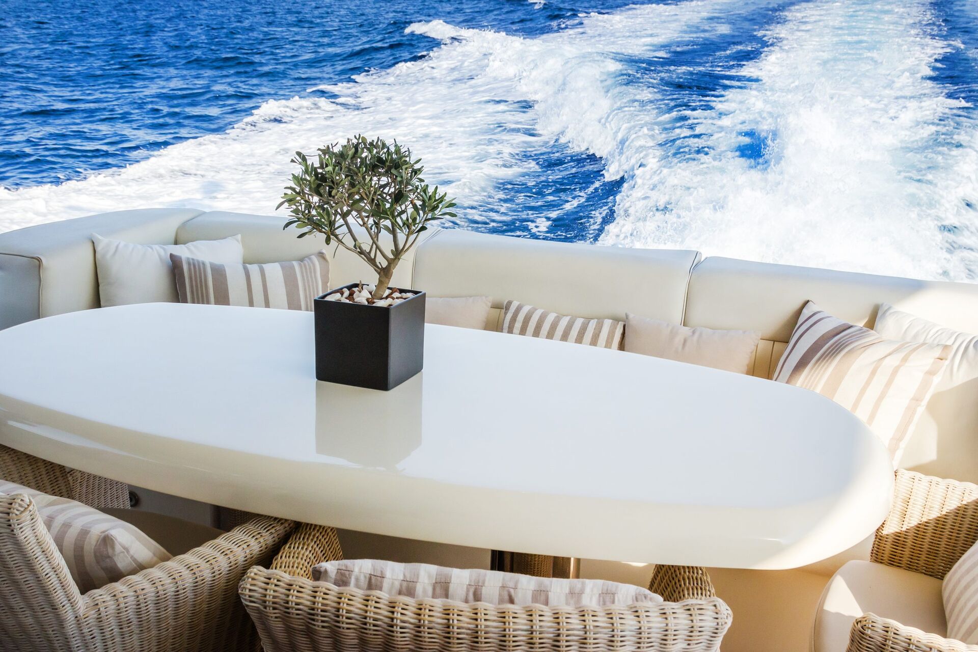 AMORAKI A white oval table with a potted plant is surrounded by wicker chairs and striped cushions on a yacht deck, with the ocean and foamy wake visible in the background.