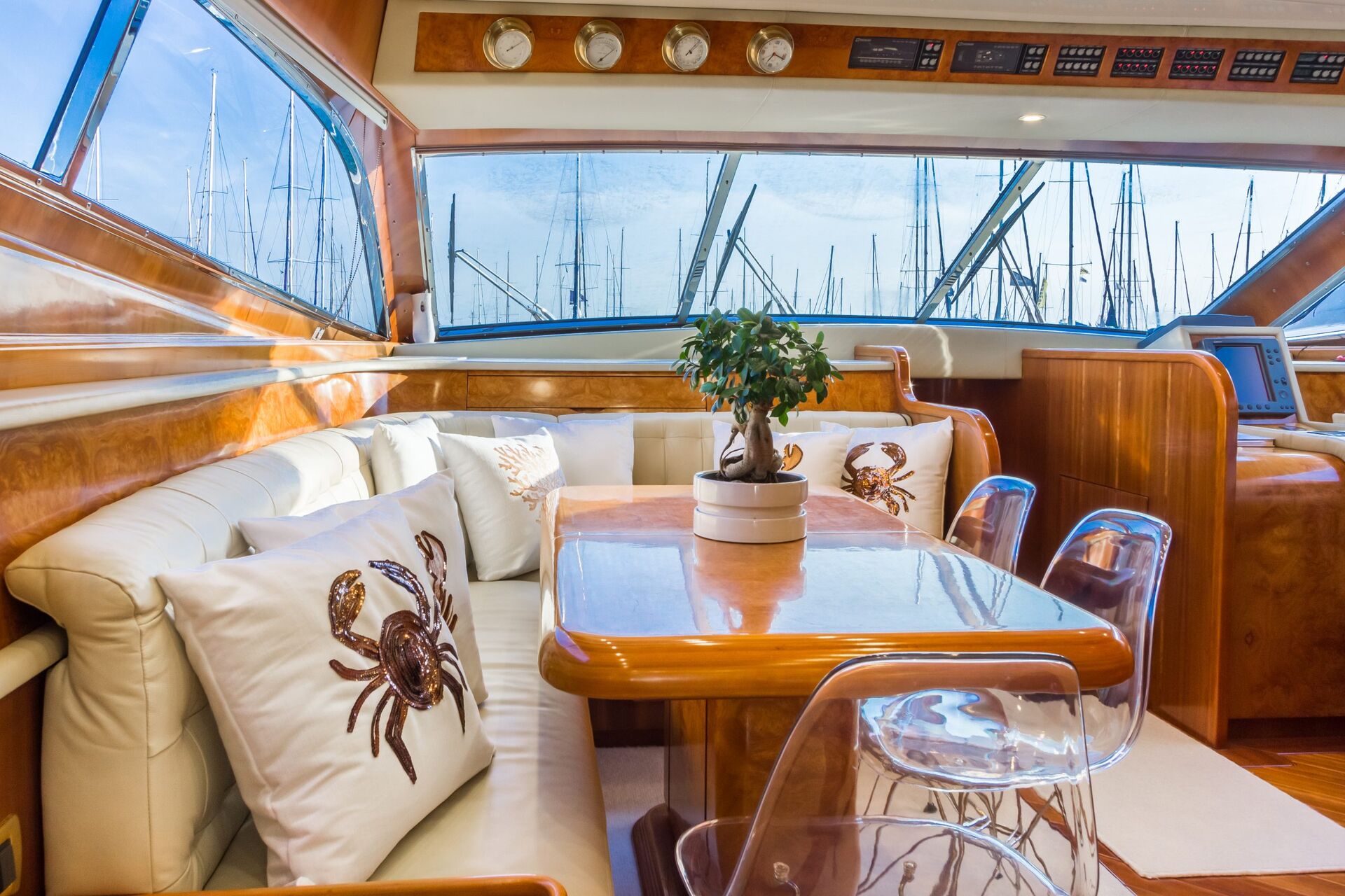 Interior of a yacht featuring a wooden table surrounded by white cushioned seating with crab-themed pillows, clear chairs, and a potted plant, with large windows overlooking a marina with sailboats.