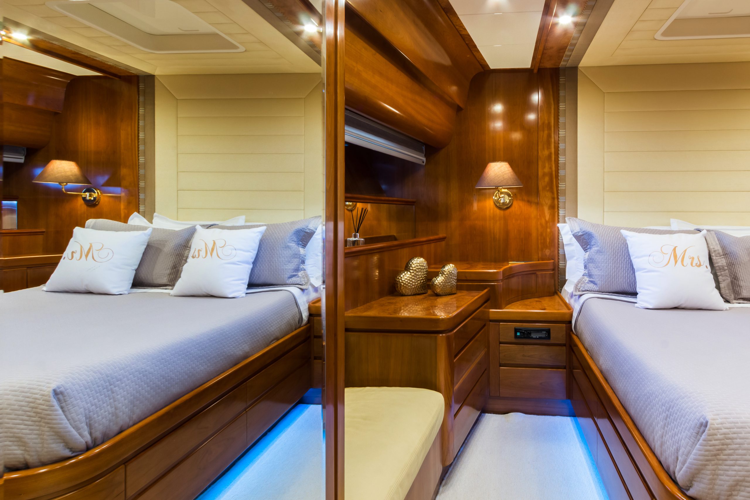 Luxurious boat cabin with wood paneling, a neatly made double bed with “Mr.” and “Mrs.” pillows, bedside lamps, a small table with decorative objects, and a mirror reflecting the room.
