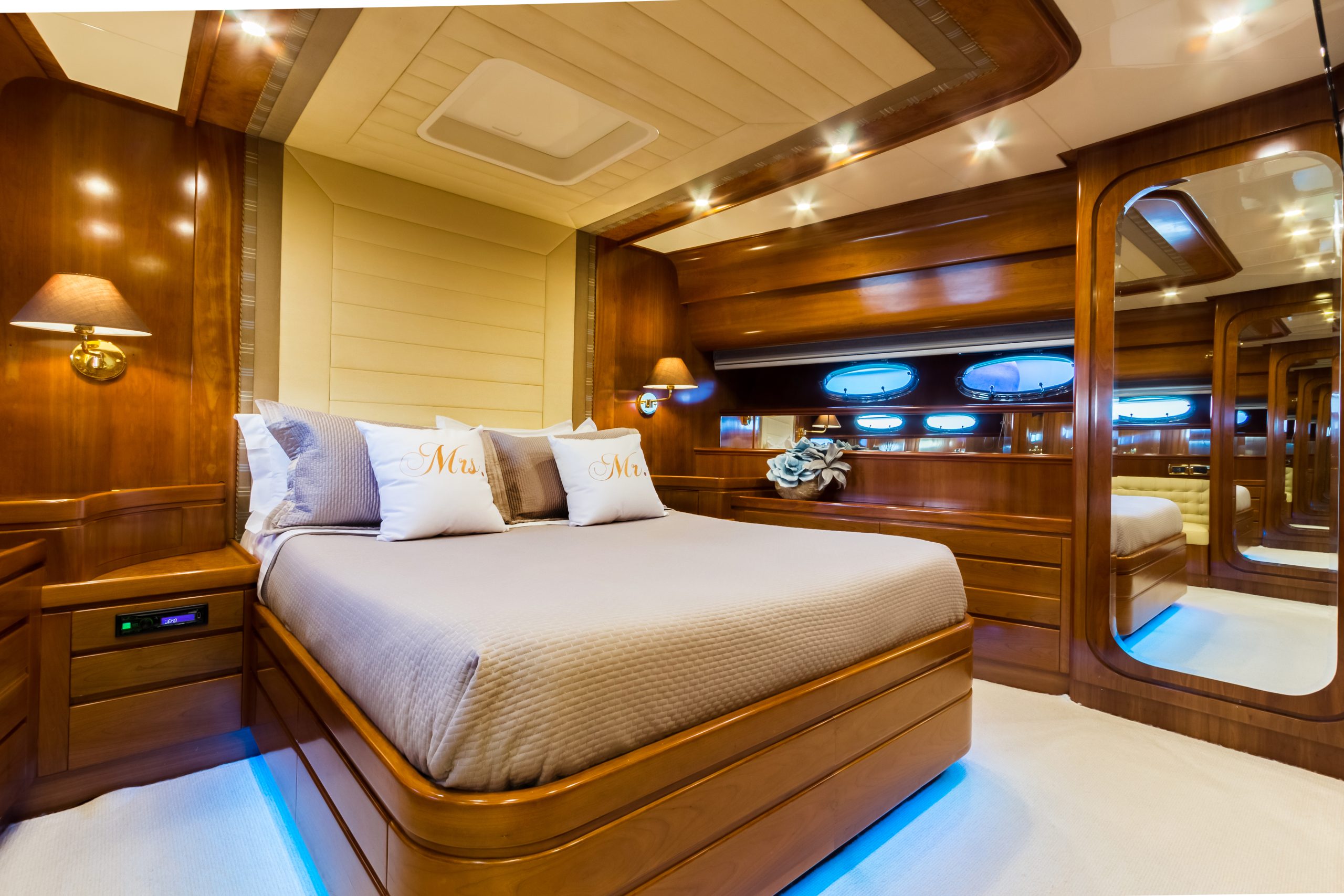 Luxurious yacht bedroom with polished wood walls, a large bed with gray bedding and “Mrs.” pillows, built-in bedside tables, round windows, ambient ceiling lights, and large mirrors reflecting the elegant space.