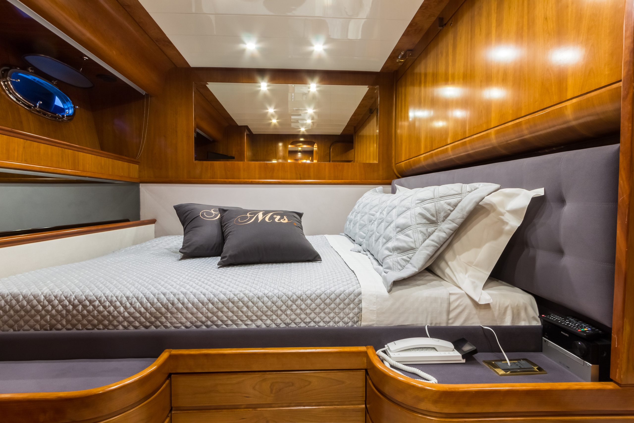 Luxurious cabin bedroom with wood paneling, a neatly made double bed with gray bedding, embroidered pillows, a wall mirror, built-in lighting, and a phone on a bedside table.