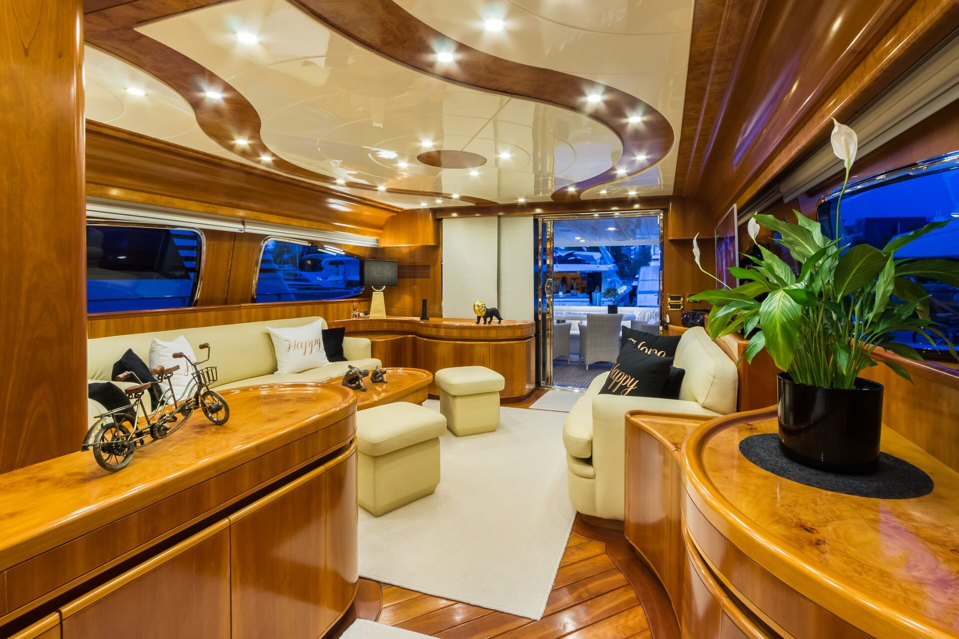 Luxurious yacht interior with polished wood finishes, cream-colored seating, decorative pillows, soft lighting, large windows, a green plant on a table, and a view of the marina through glass doors.