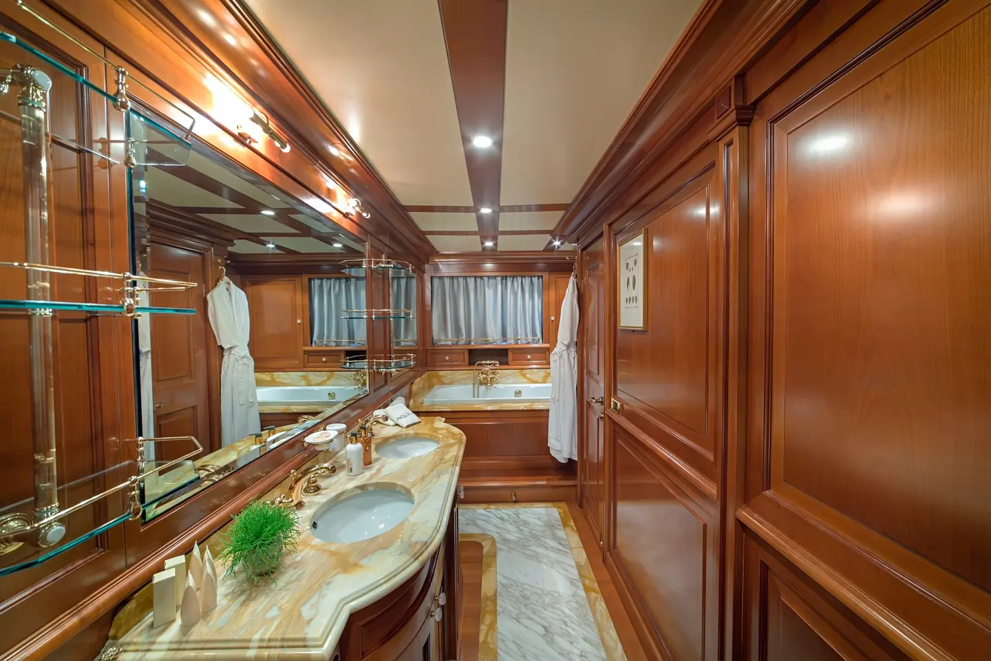 Luxurious bathroom with wood-paneled walls, marble countertop with double sinks, mirrors, shelves with toiletries, white robes, and bright lighting.