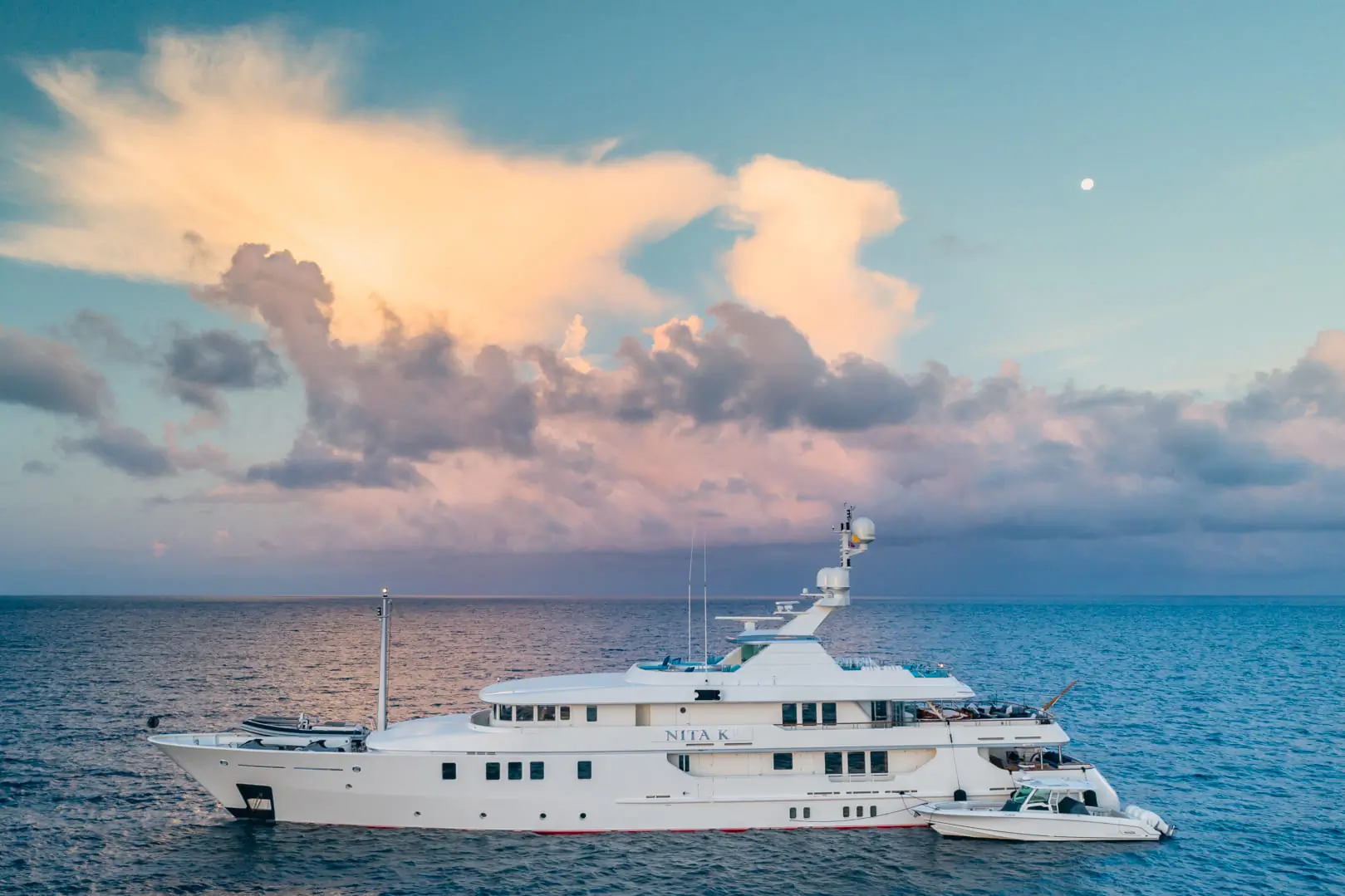 A large white yacht floats on calm blue water at sunset, with a small boat beside it. Pink and orange clouds fill the sky, and the moon is visible above the horizon.