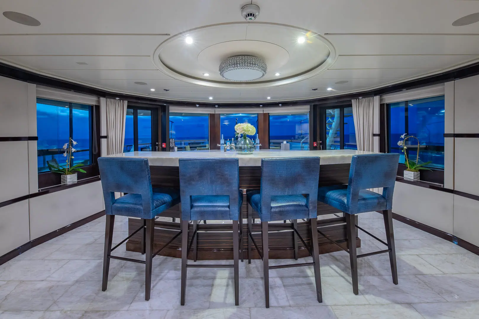 A modern indoor bar area with five blue cushioned chairs, a round counter, white marble floor, large windows showing an ocean view, and a decorative ceiling light.