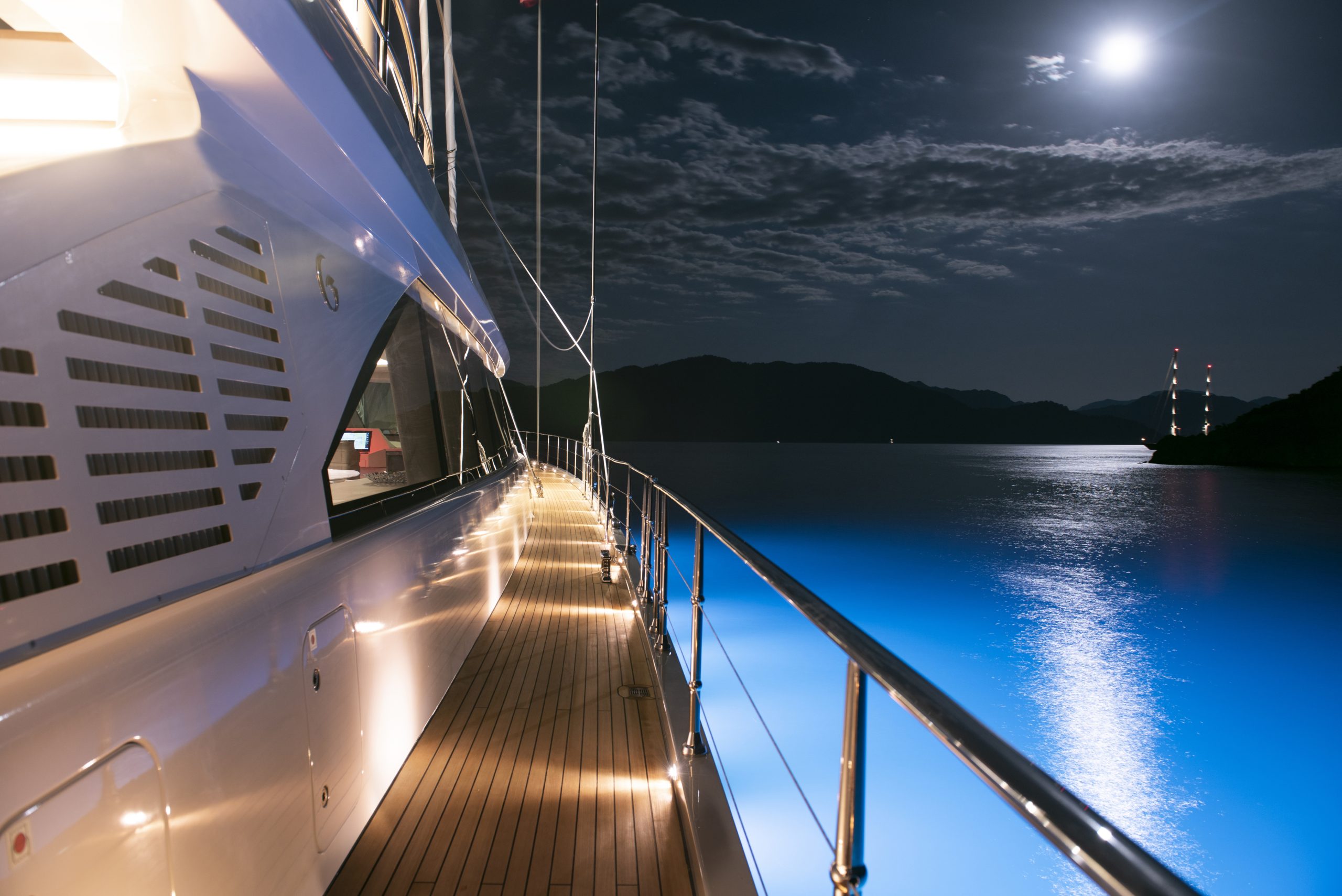 A yacht deck illuminated at night, with glowing blue water below, mountains in the distance, and a bright full moon shining in a partly cloudy sky.
