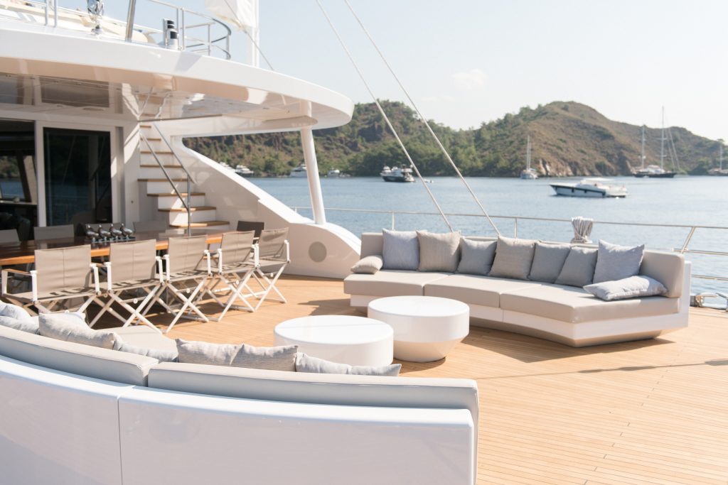 Spacious yacht deck with modern white sofas, a dining table with chairs, and round tables. Calm water and several boats docked near a hilly shoreline are visible in the background under a clear sky.