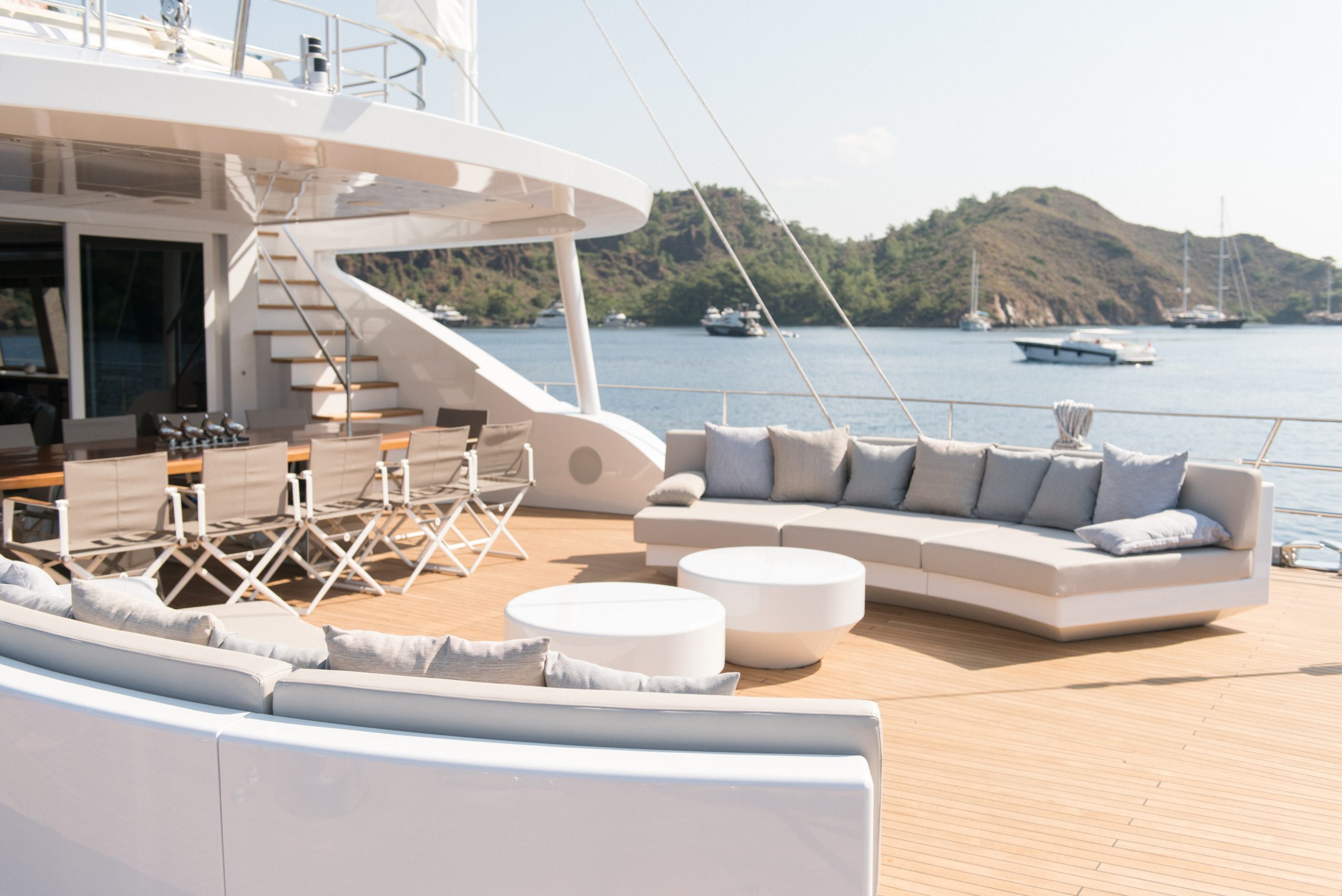 Spacious yacht deck with modern white sofas, a dining table with chairs, and round tables. Calm water and several boats docked near a hilly shoreline are visible in the background under a clear sky.