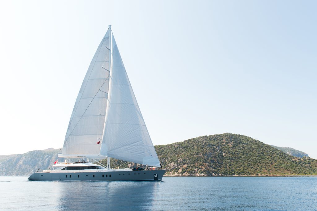 A large white sailboat with tall sails glides on calm blue water near a green, hilly coastline under a clear sky.