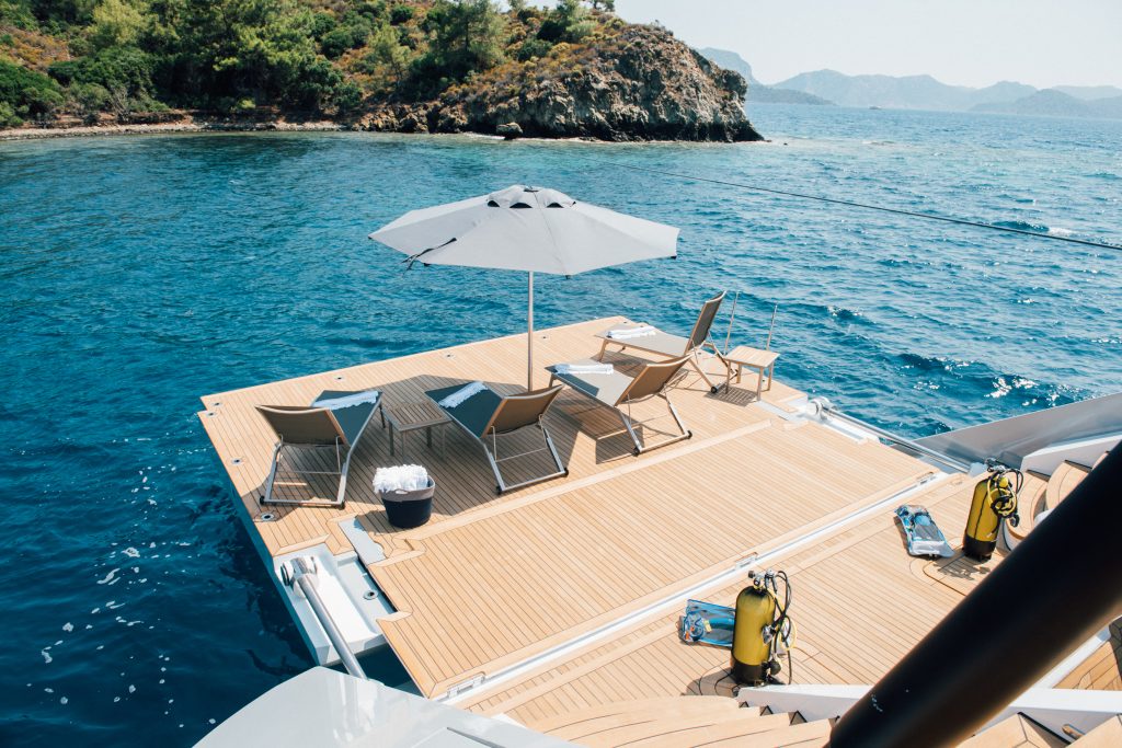 A spacious yacht deck with lounge chairs and an umbrella overlooks clear blue ocean water. Scuba tanks are nearby, and a rocky, green island is in the background under a sunny sky.
