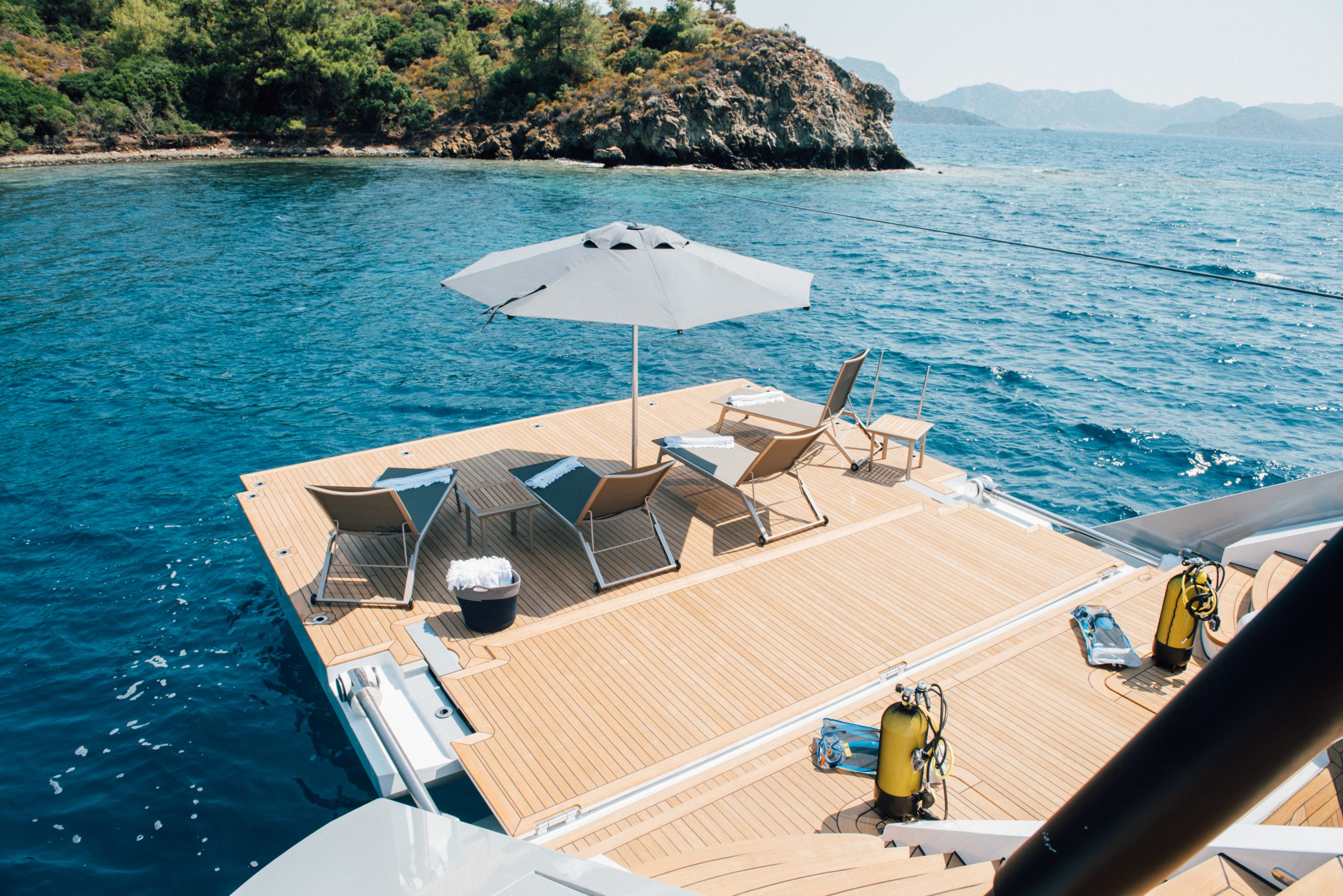 A spacious yacht deck with lounge chairs and an umbrella overlooks clear blue ocean water. Scuba tanks are nearby, and a rocky, green island is in the background under a sunny sky.