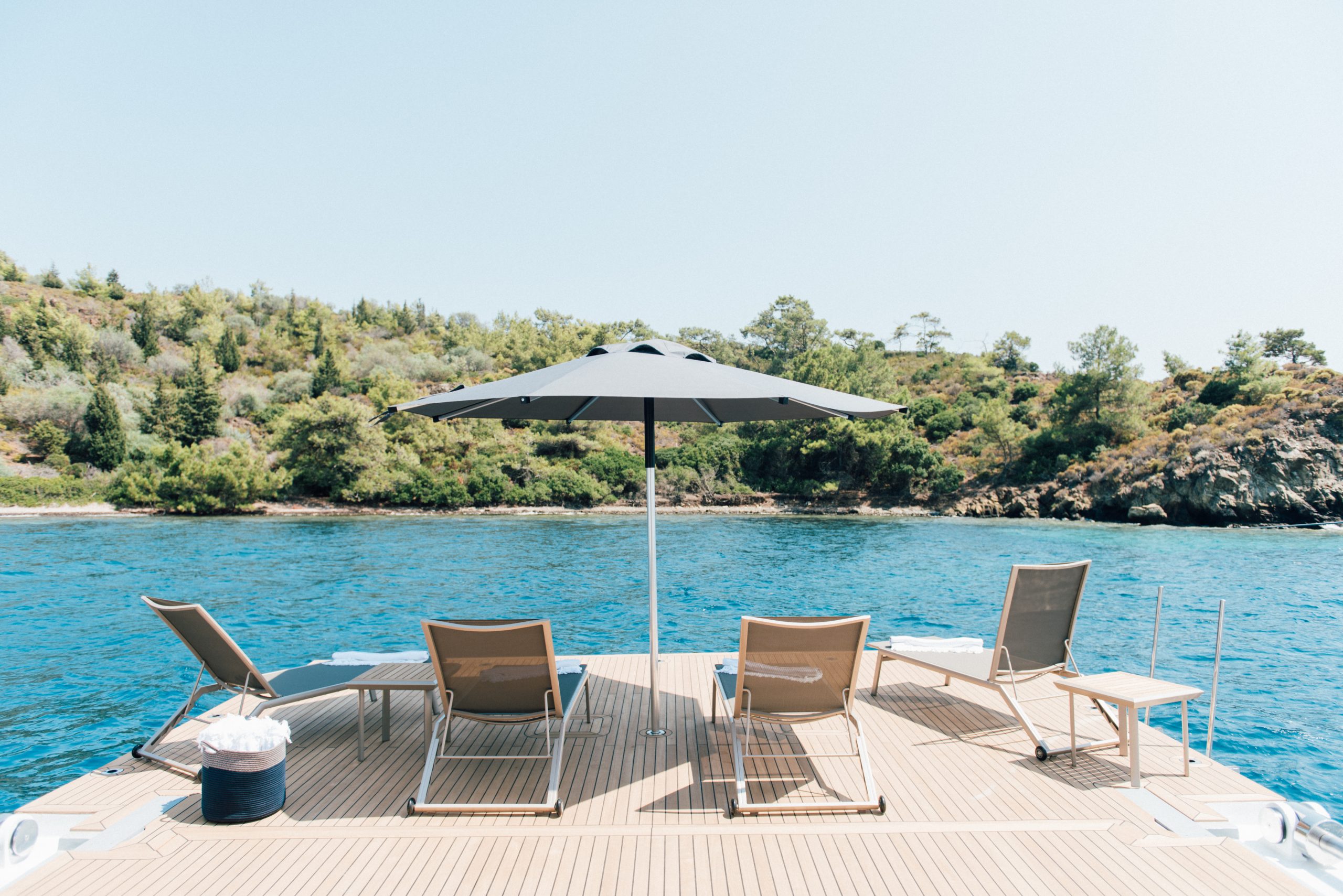 Four lounge chairs and a large umbrella on a wooden dock overlook calm blue water and a forested shoreline under a clear sky, creating a relaxing and scenic setting.