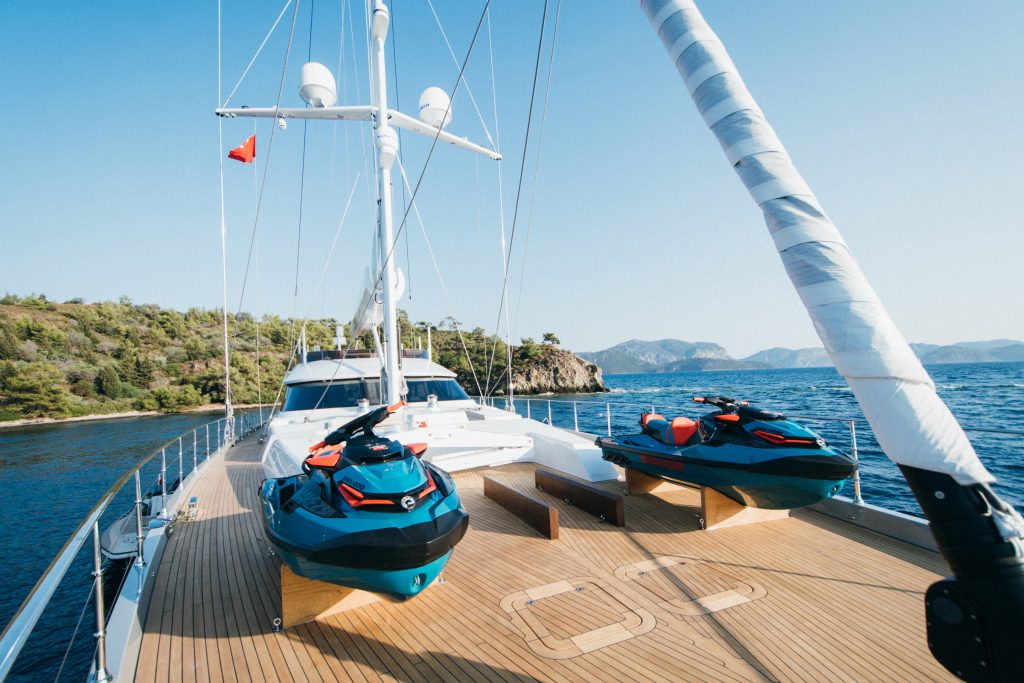 A luxury yacht deck with two jet skis secured at the front, surrounded by calm blue water and distant green hills under a clear sky.