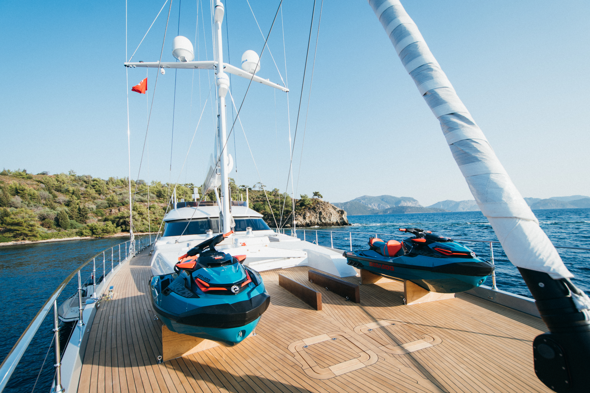 A luxury yacht deck with two jet skis secured at the front, surrounded by calm blue water and distant green hills under a clear sky.