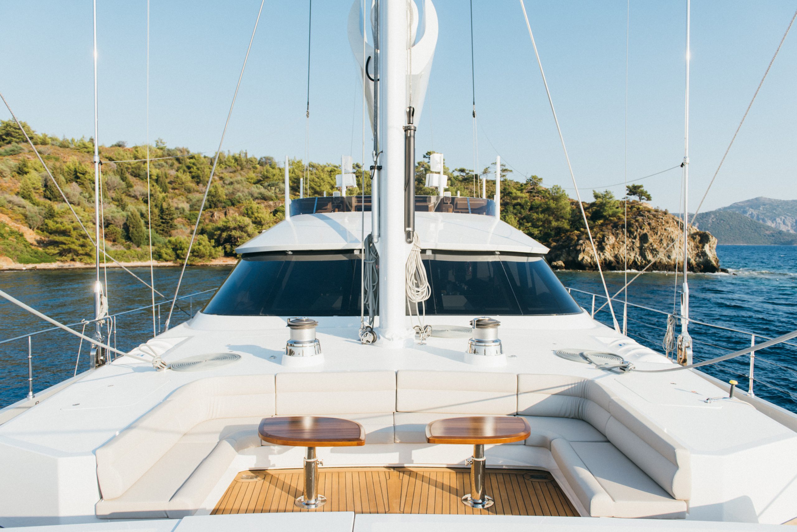 View from a yacht’s deck showing white cushioned seating and two wooden tables, with calm blue water and a scenic, tree-covered coastline in the background under a clear sky.