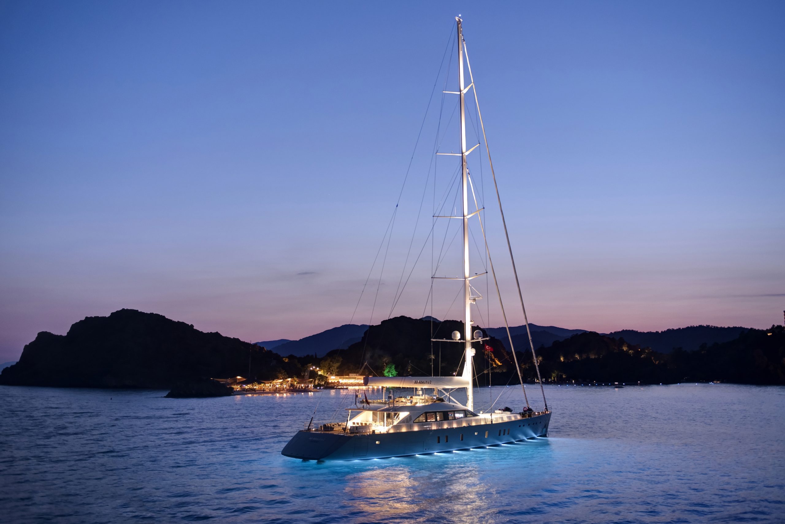 A luxury sailboat with illuminated decks is anchored on calm water at dusk, with coastal hills and distant lights visible against a twilight sky.
