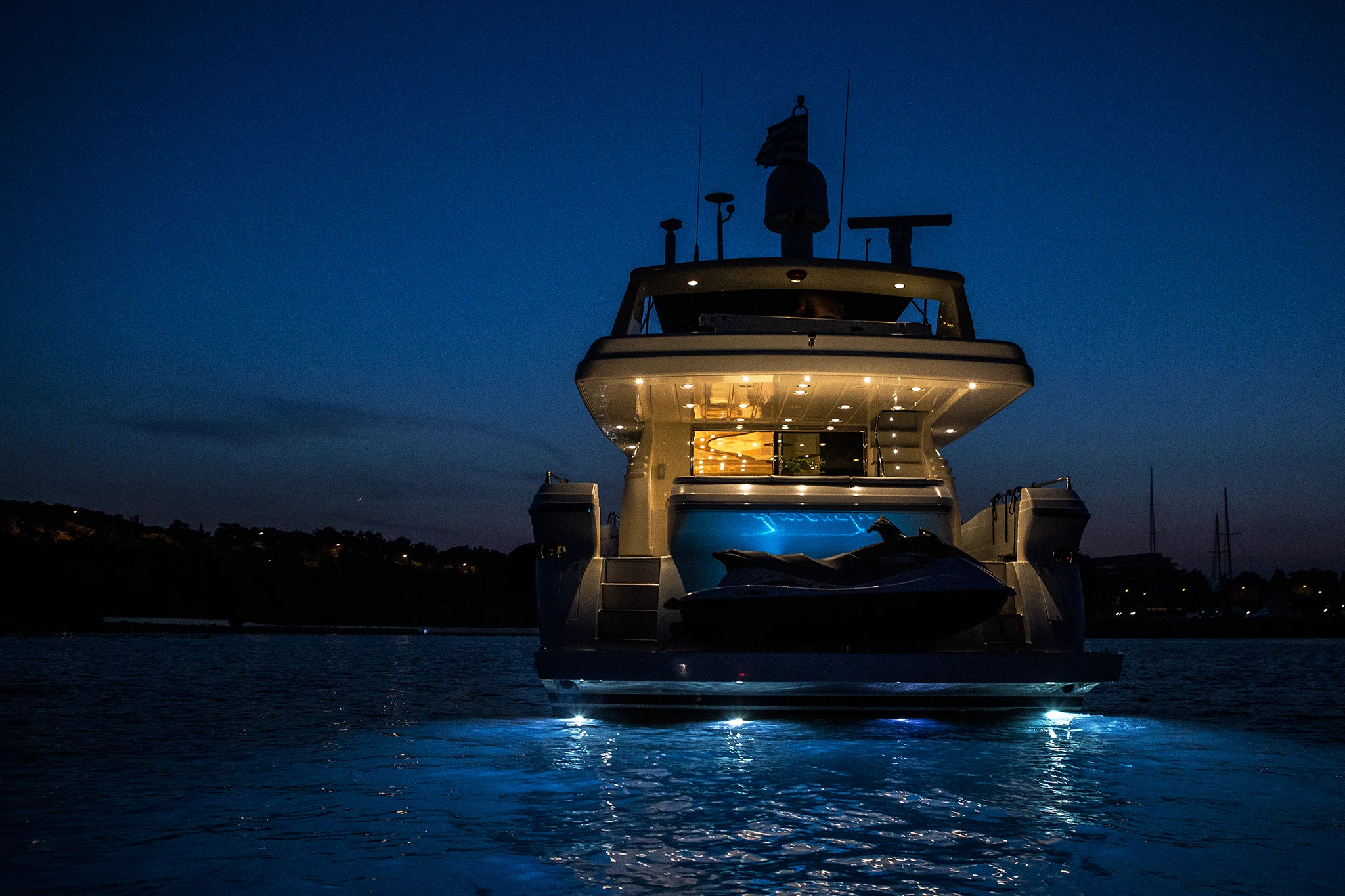 A luxury yacht illuminated with blue underwater lights is anchored on calm water at dusk, with its interior lights glowing and a jet ski visible at the back. Distant shoreline and a dark blue sky complete the scene.