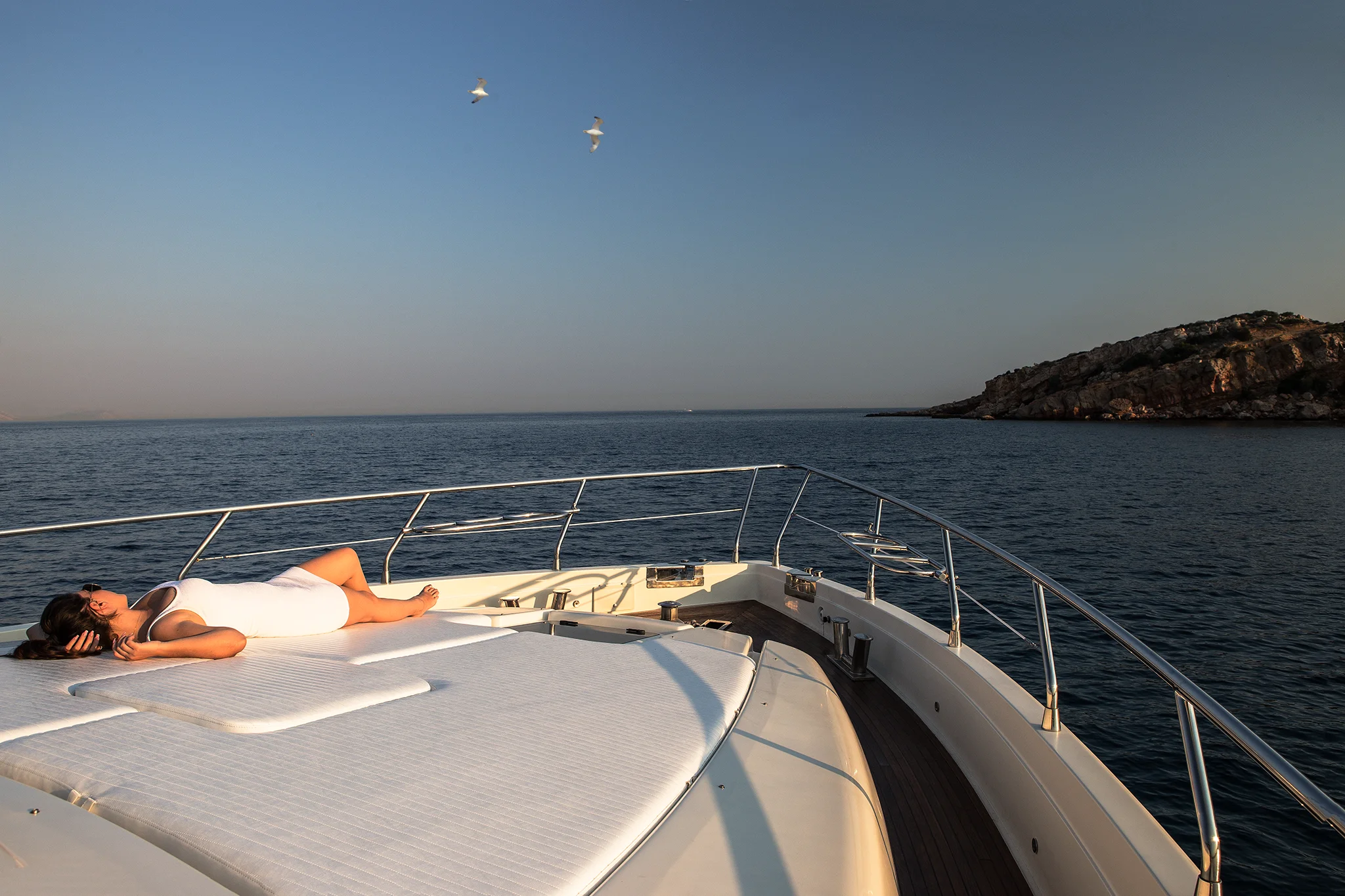A woman in a white dress relaxes on the deck of a yacht, basking in the sun. The yacht is on calm blue water near a rocky shore, with a clear sky and three birds flying overhead.