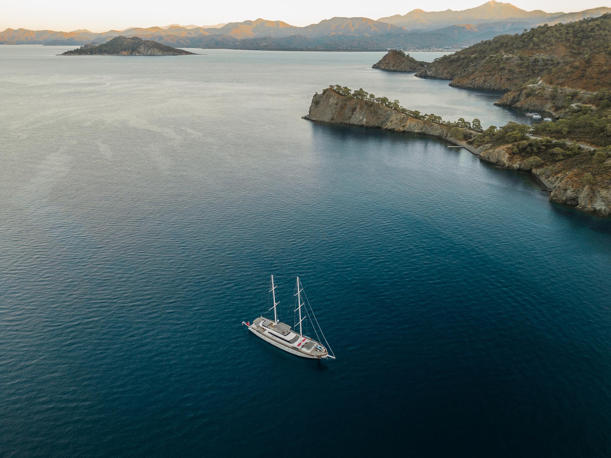 A white sailboat floats alone on calm blue water near rocky green cliffs, with distant mountains under a hazy sky in the background.