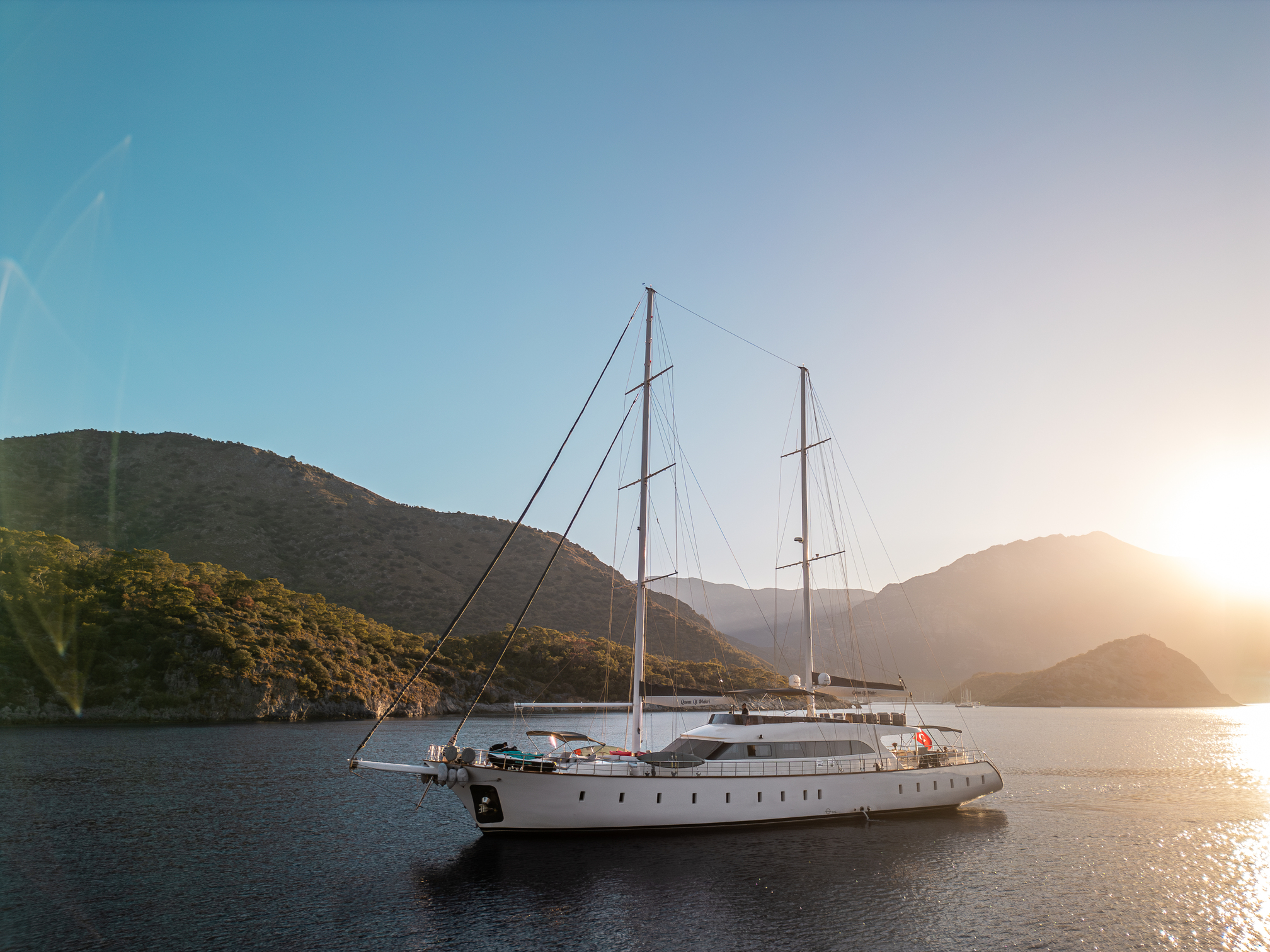 A large white yacht is anchored on calm water near a mountainous coastline at sunset, with the sun low in the sky and hills silhouetted in the background.