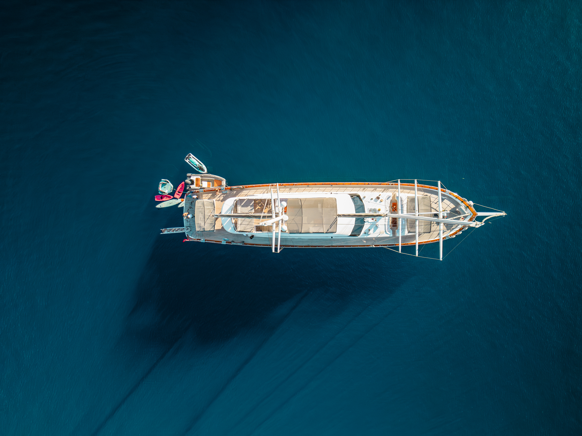 Aerial view of a yacht anchored in deep blue water, with paddleboards and a small boat attached at the stern. The sunlight highlights the yacht’s deck and creates shadows on the calm, clear sea.