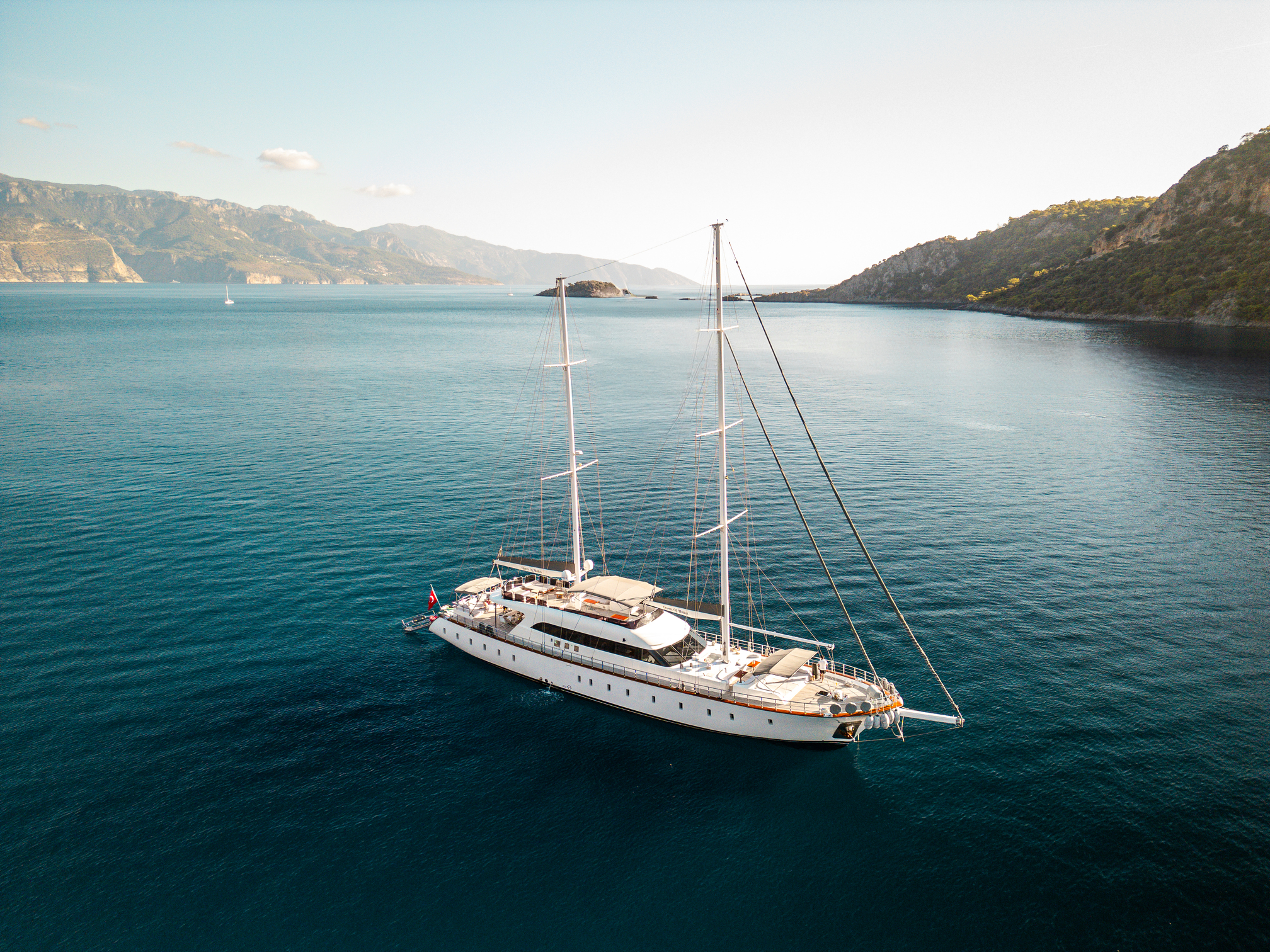 A large white sailing yacht with three masts floats on calm blue water, surrounded by coastal mountains and cliffs under a clear sky.