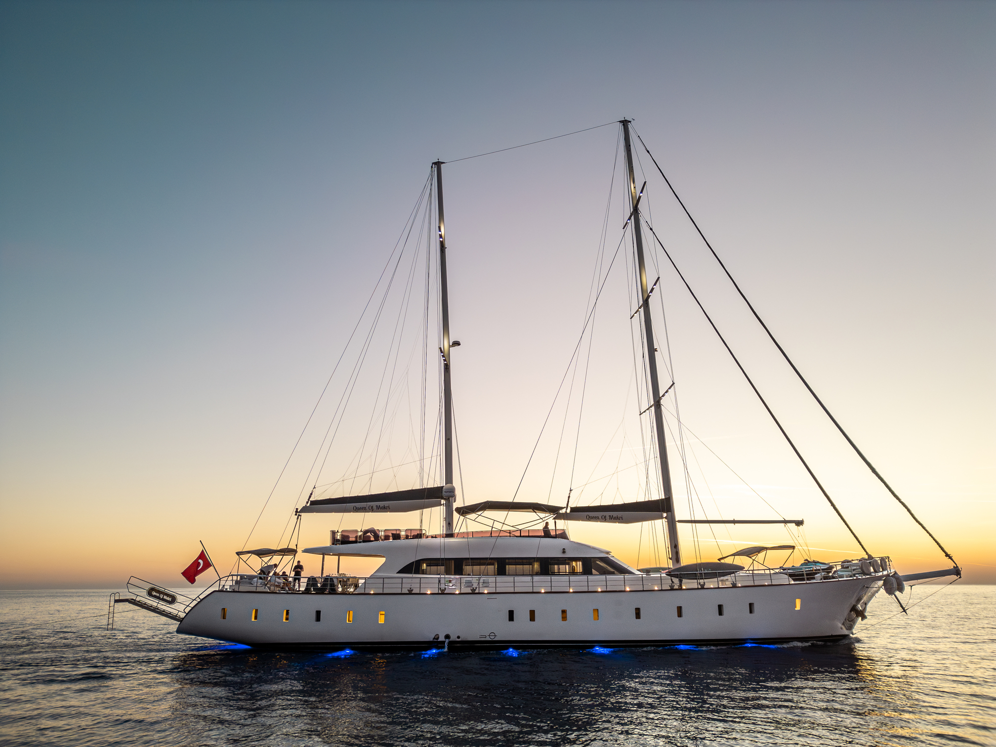 A large, modern white sailing yacht is anchored on calm water at sunset, with exterior lights glowing and a red flag at the stern. The sky is clear with warm colors near the horizon.