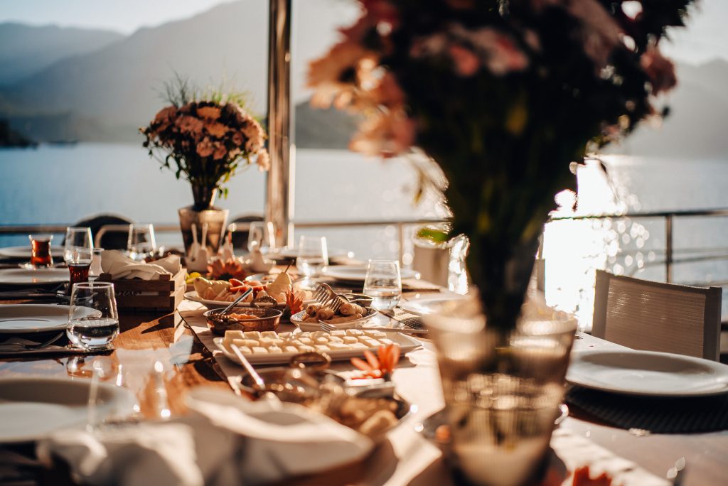 A sunlit outdoor dining table set with plates, glasses, flowers, and an assortment of food, overlooking a lake and mountains in the background.