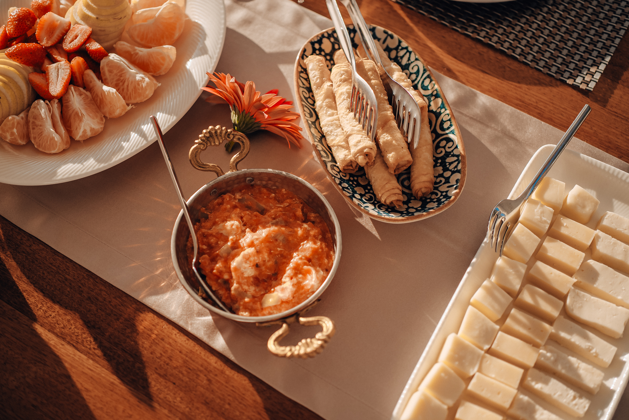 A table set with plates of sliced cheese, rolled pastries, a tomato-based dip in a small pot, peeled mandarin segments, and strawberries, with a flower for decoration and warm sunlight illuminating the scene.
