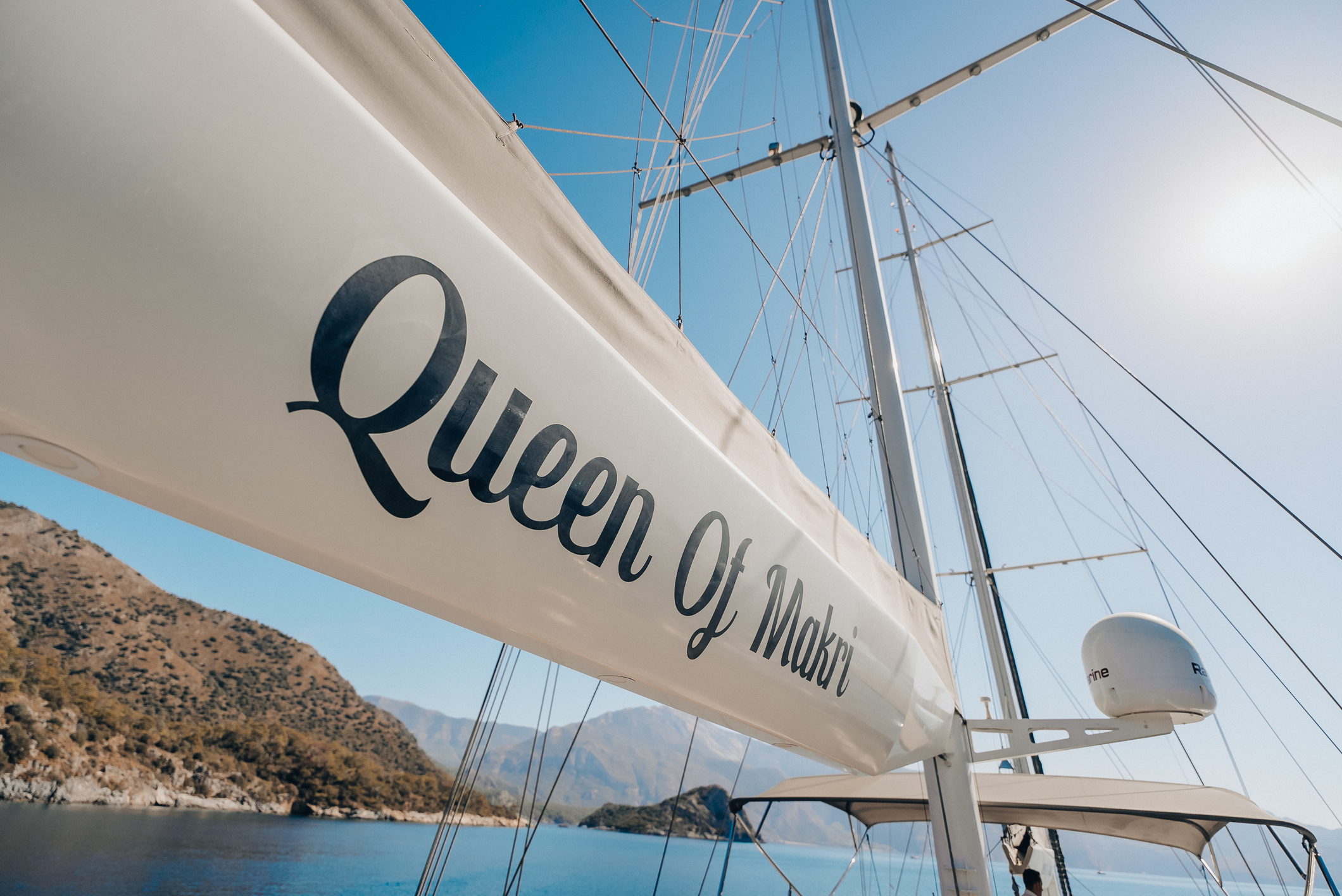 Close-up view of a sailboat with the name Queen Of Makri written on the sail. The boat is on calm blue water near a mountainous coastline under a clear, sunny sky.