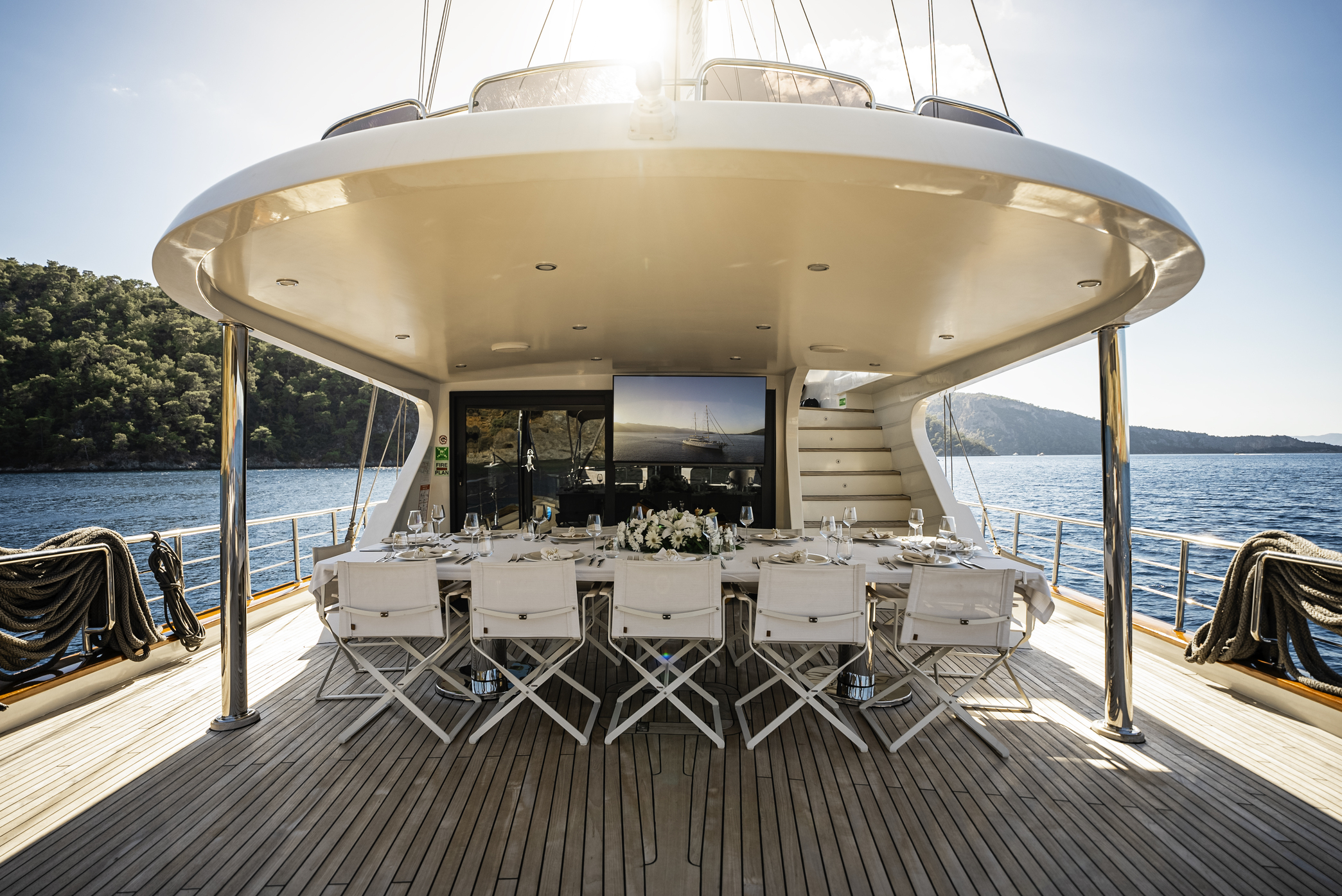 Luxurious outdoor dining area on the deck of a yacht, set with a long table, white chairs, floral centerpiece, and views of blue water and green hills in the background under a sunny sky.