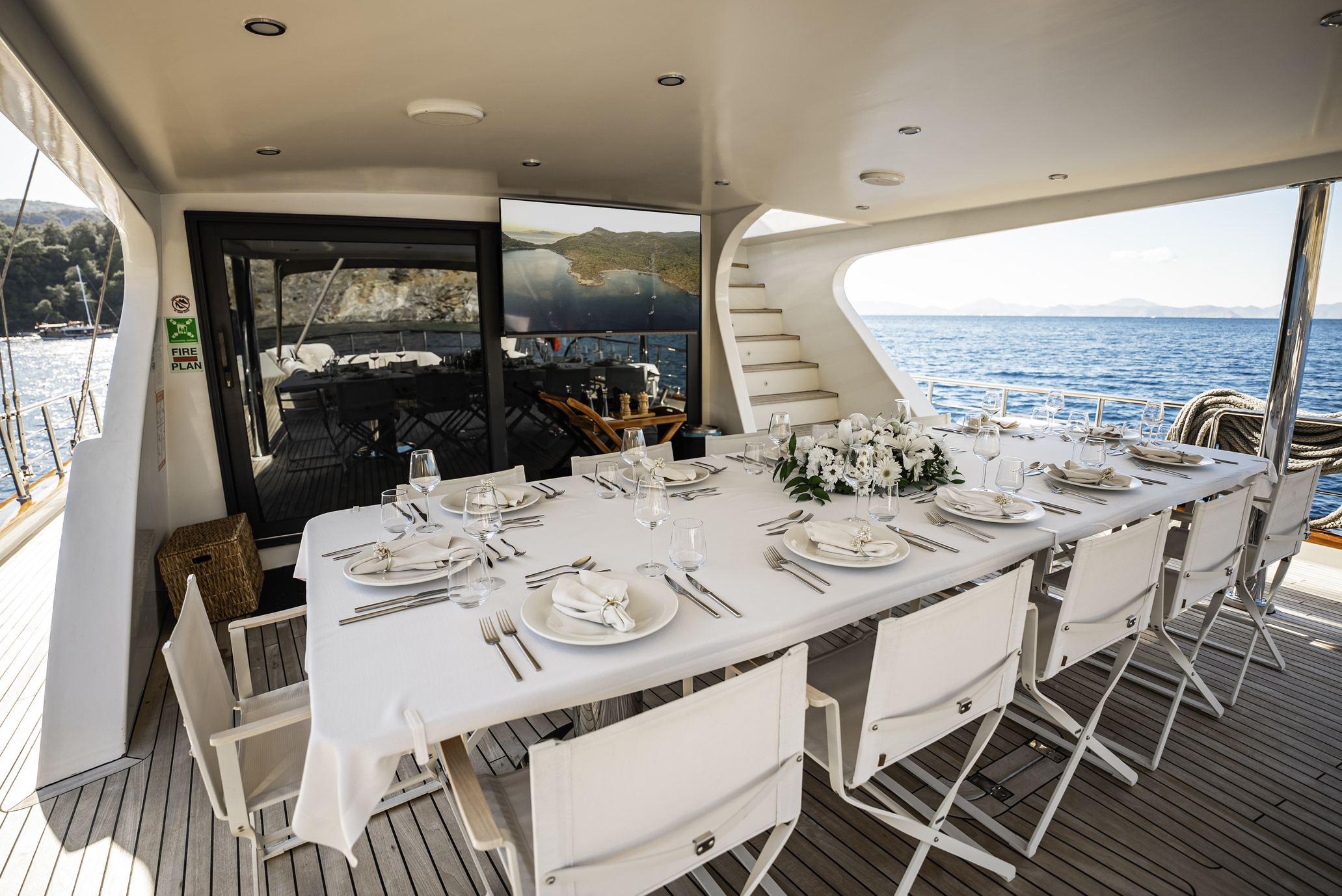 A yacht deck set for dining with a long white table, elegant place settings, white chairs, and a centerpiece of white flowers. The sea and distant hills are visible in the background under a sunny sky.