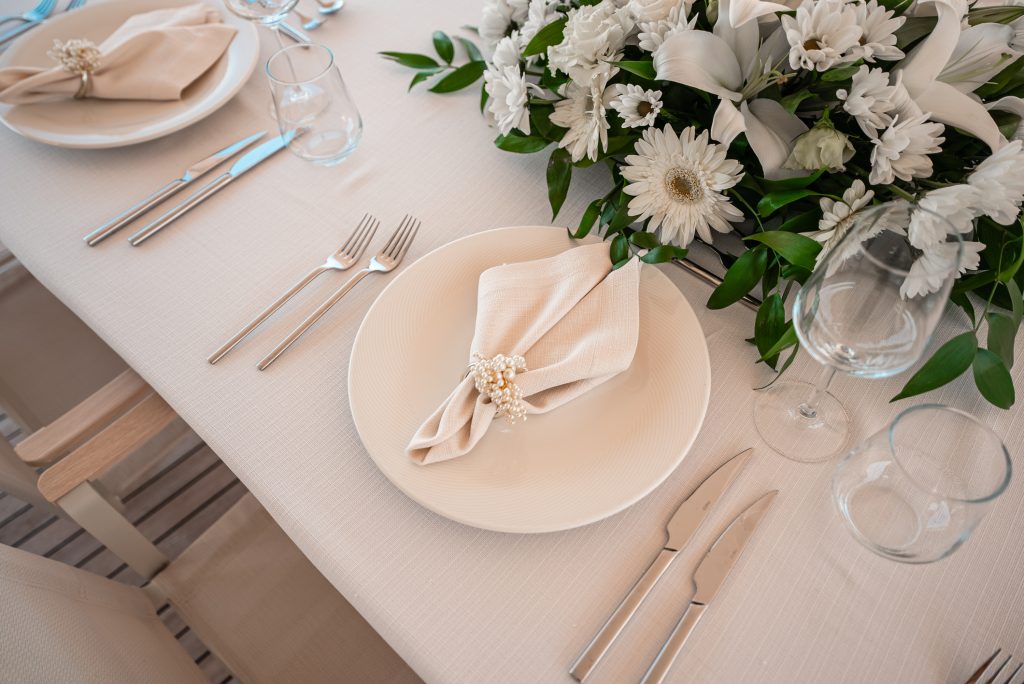 An elegant table setting with a white plate, beige napkin, and pearl napkin ring, surrounded by silver cutlery, glassware, and a floral centerpiece with white lilies and daisies on a cream tablecloth.