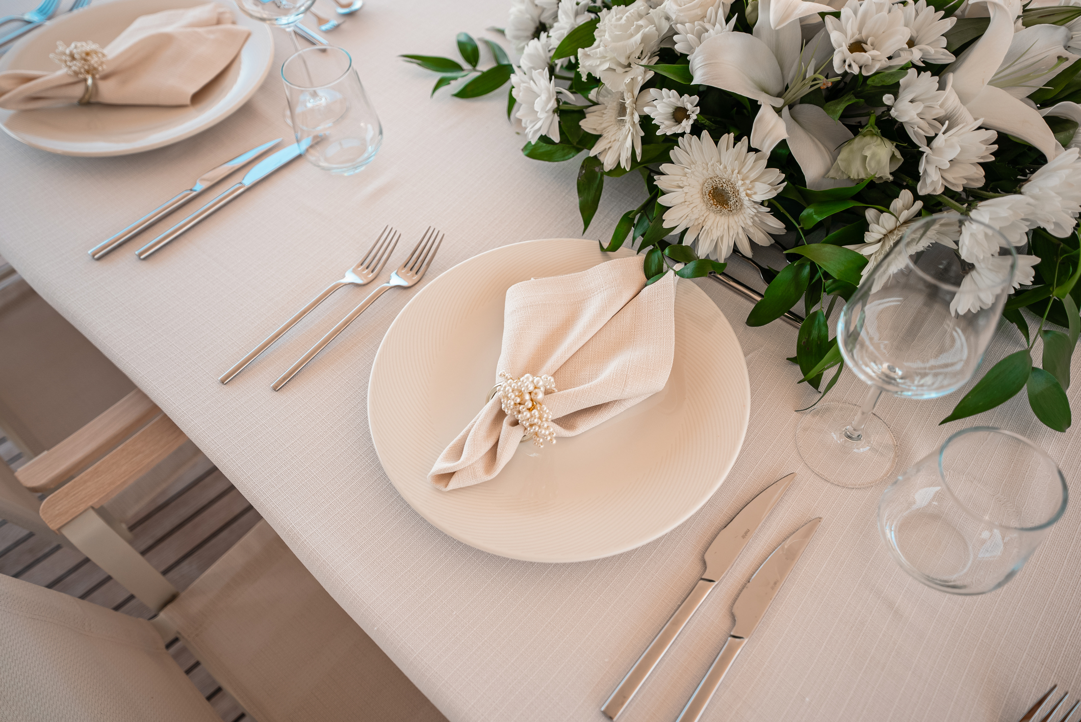 An elegant table setting with a white plate, beige napkin, and pearl napkin ring, surrounded by silver cutlery, glassware, and a floral centerpiece with white lilies and daisies on a cream tablecloth.