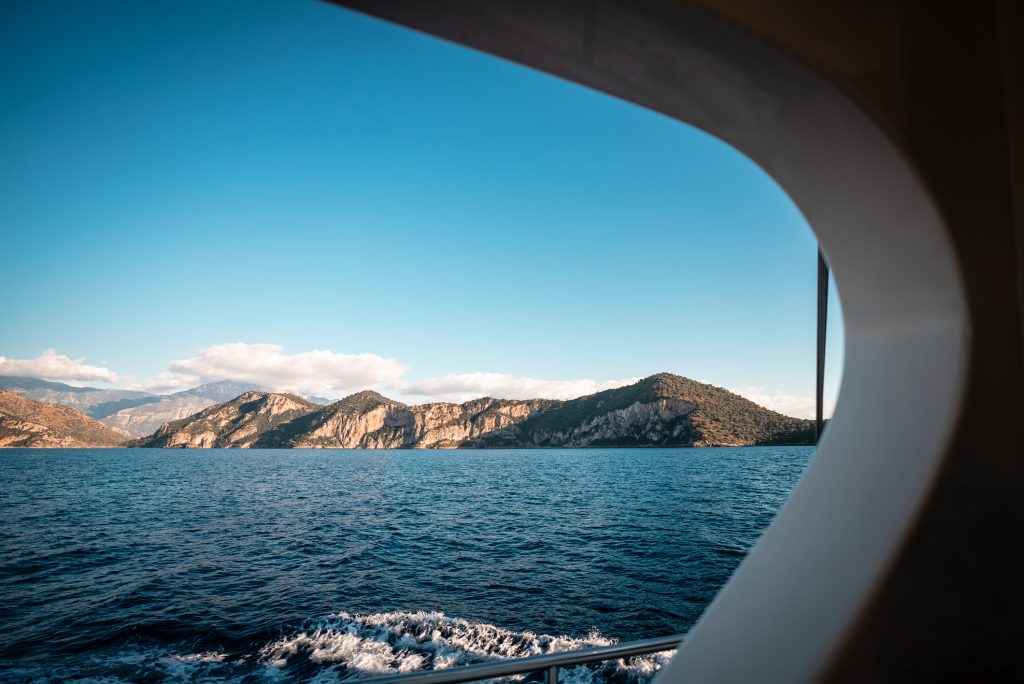 View of a blue sea and distant green mountains, seen through the curved window frame of a boat under a clear sky with a few clouds.